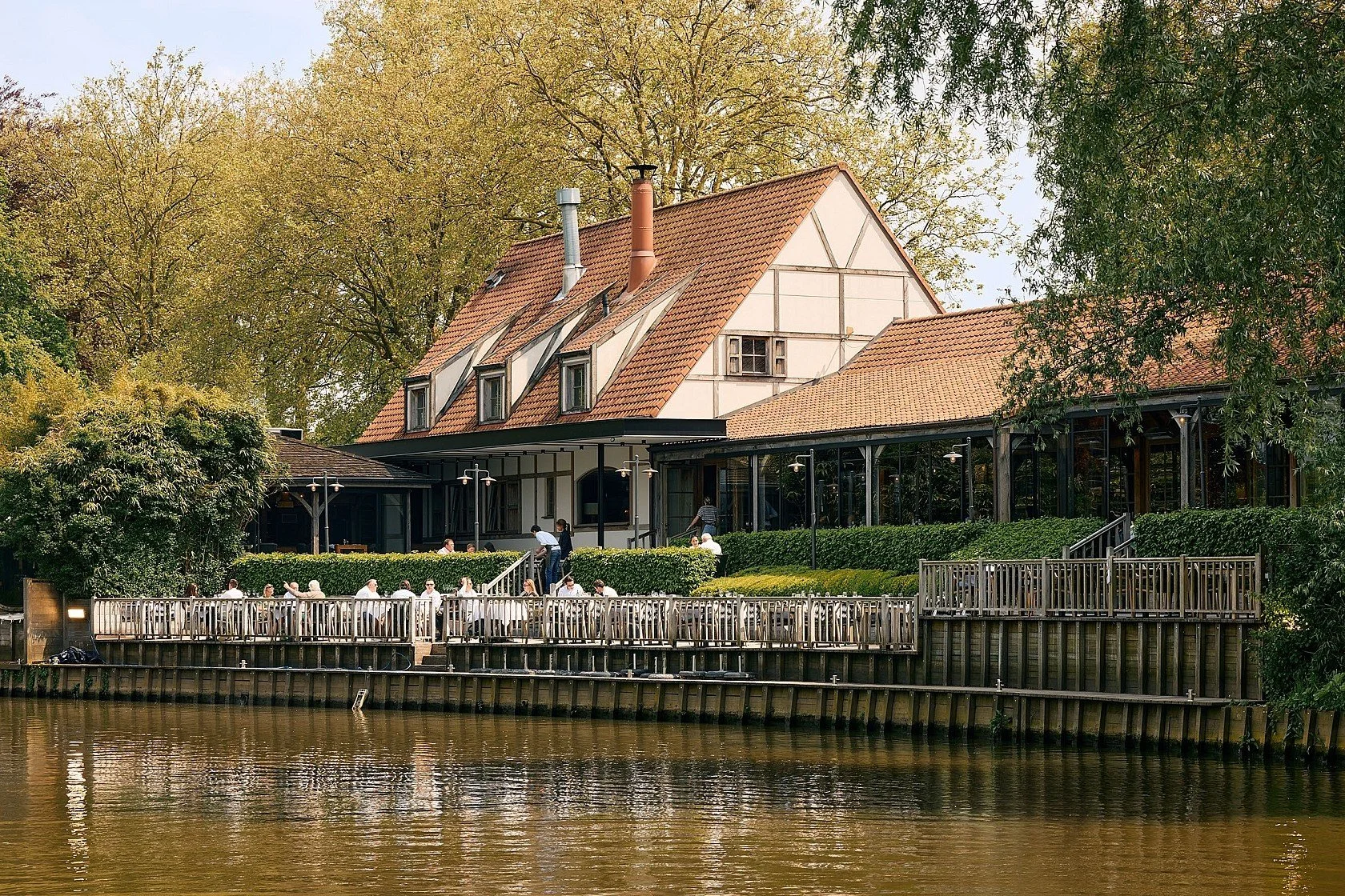 Een restauranthuis gelegen aan een rivier met een houten terras en mensen die daar eten en drinken, omringd door groene bomen.