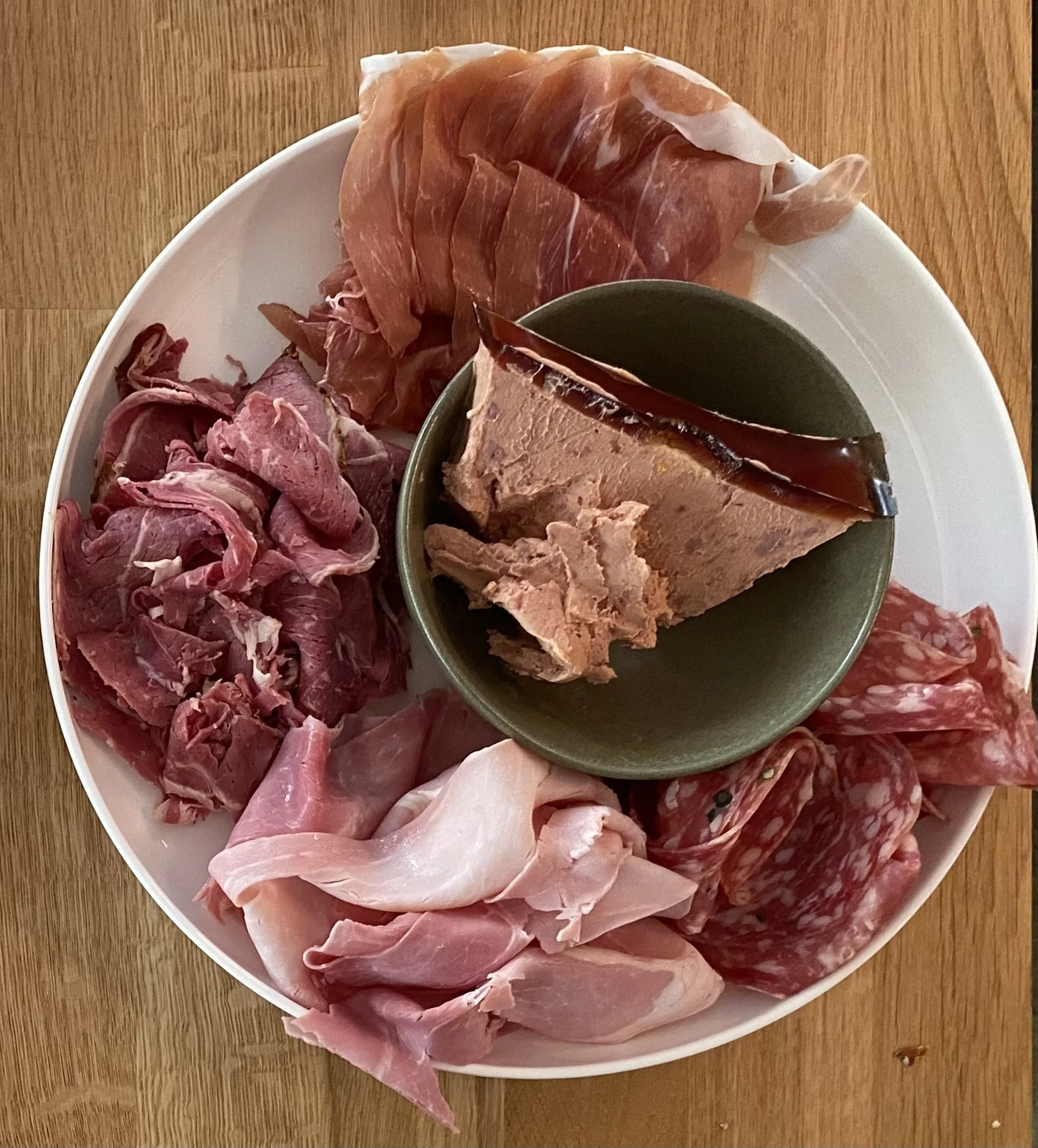 Een grote witte schaal met diverse soorten rauwe vleeswaren en een klein schaaltje met gelei of mousse in het midden.