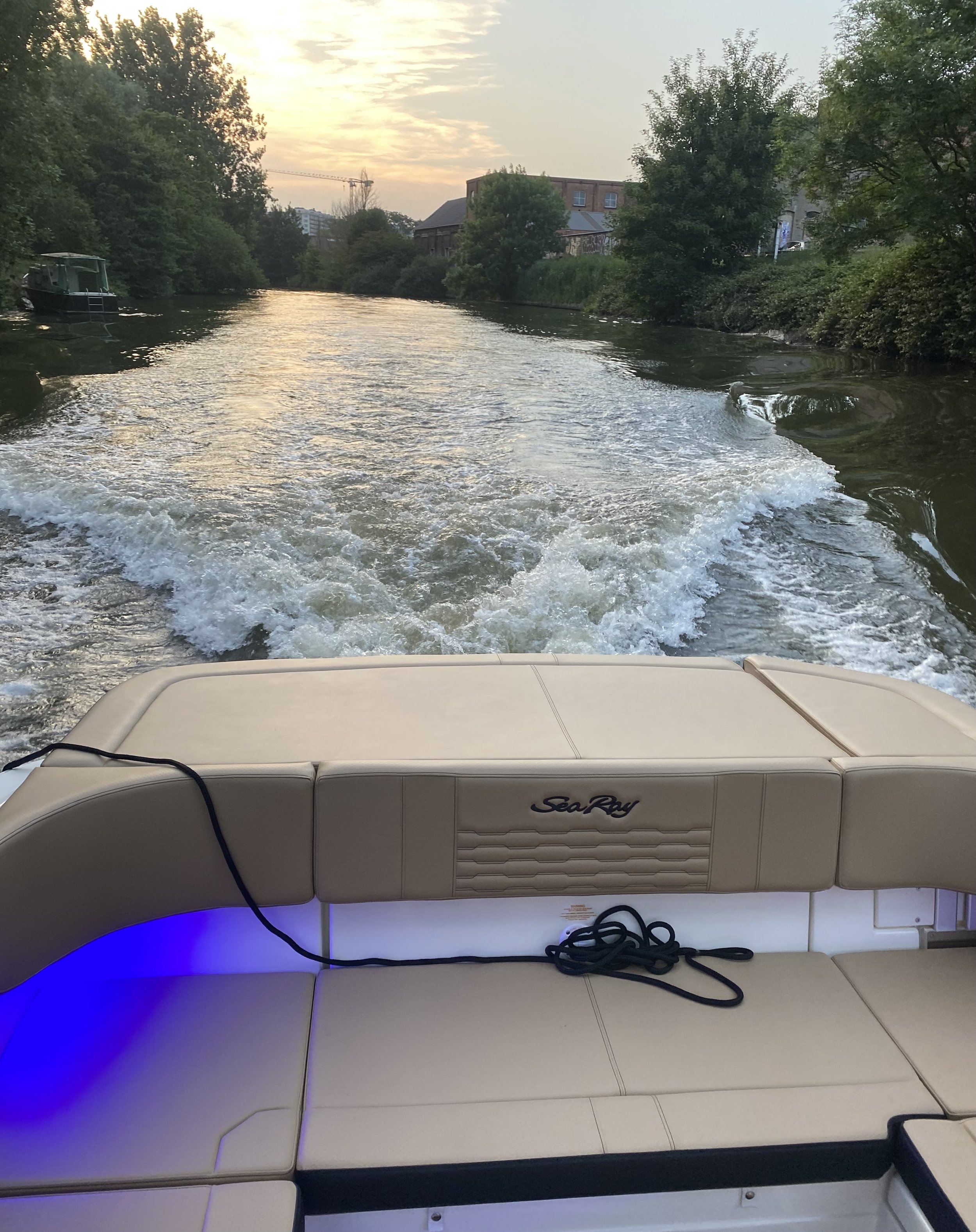 Op de afbeelding is de achterkant van een wit boot te zien die over een rivier vaart, met de roeispanen achter zich en een kalme rivier met bomen en gebouwen op de oevers.