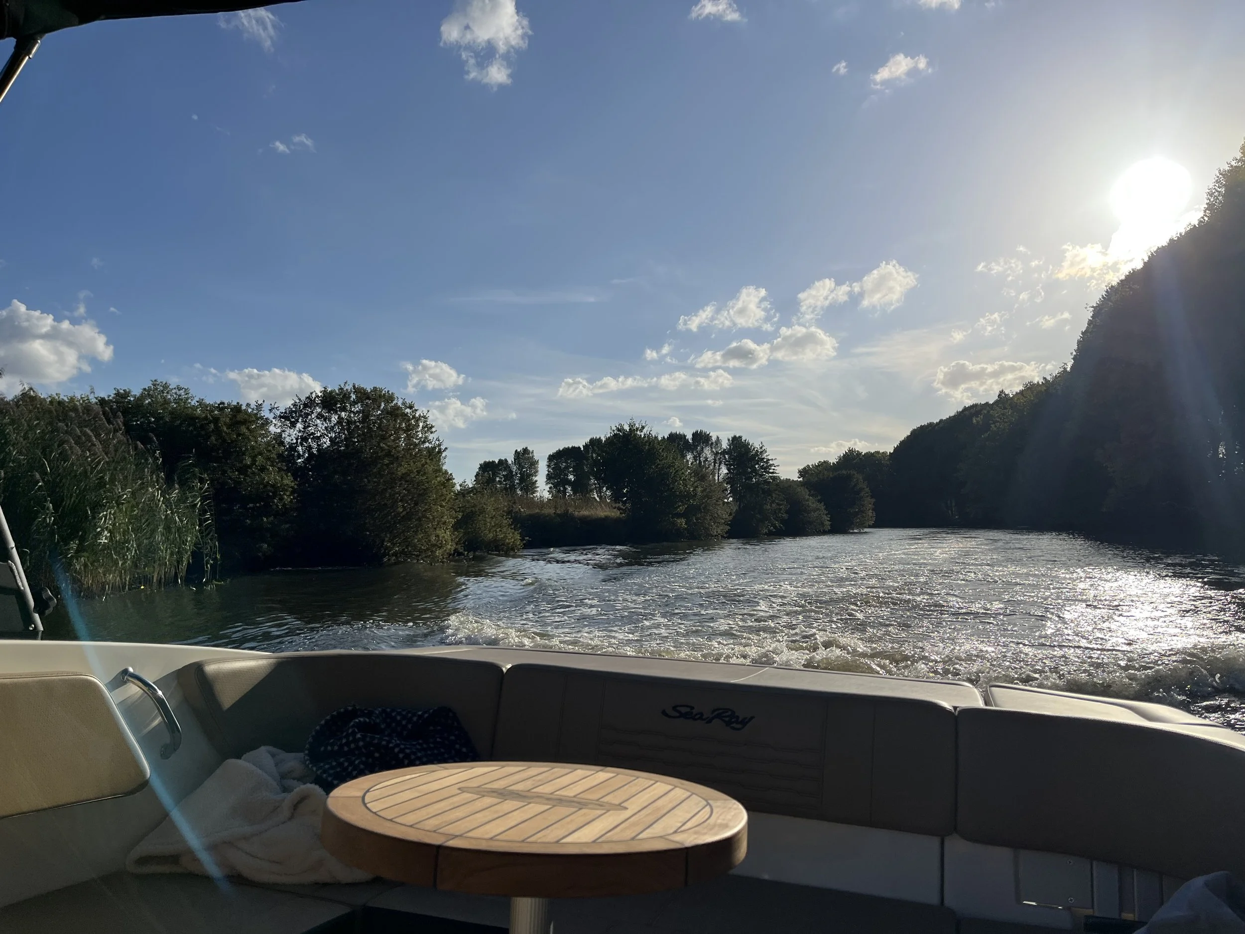 Uitzicht vanaf een boot over een rivier met groene bomen langs de oevers, onder een blauwe hemel met enkele witte wolken en de zon die schijnt.