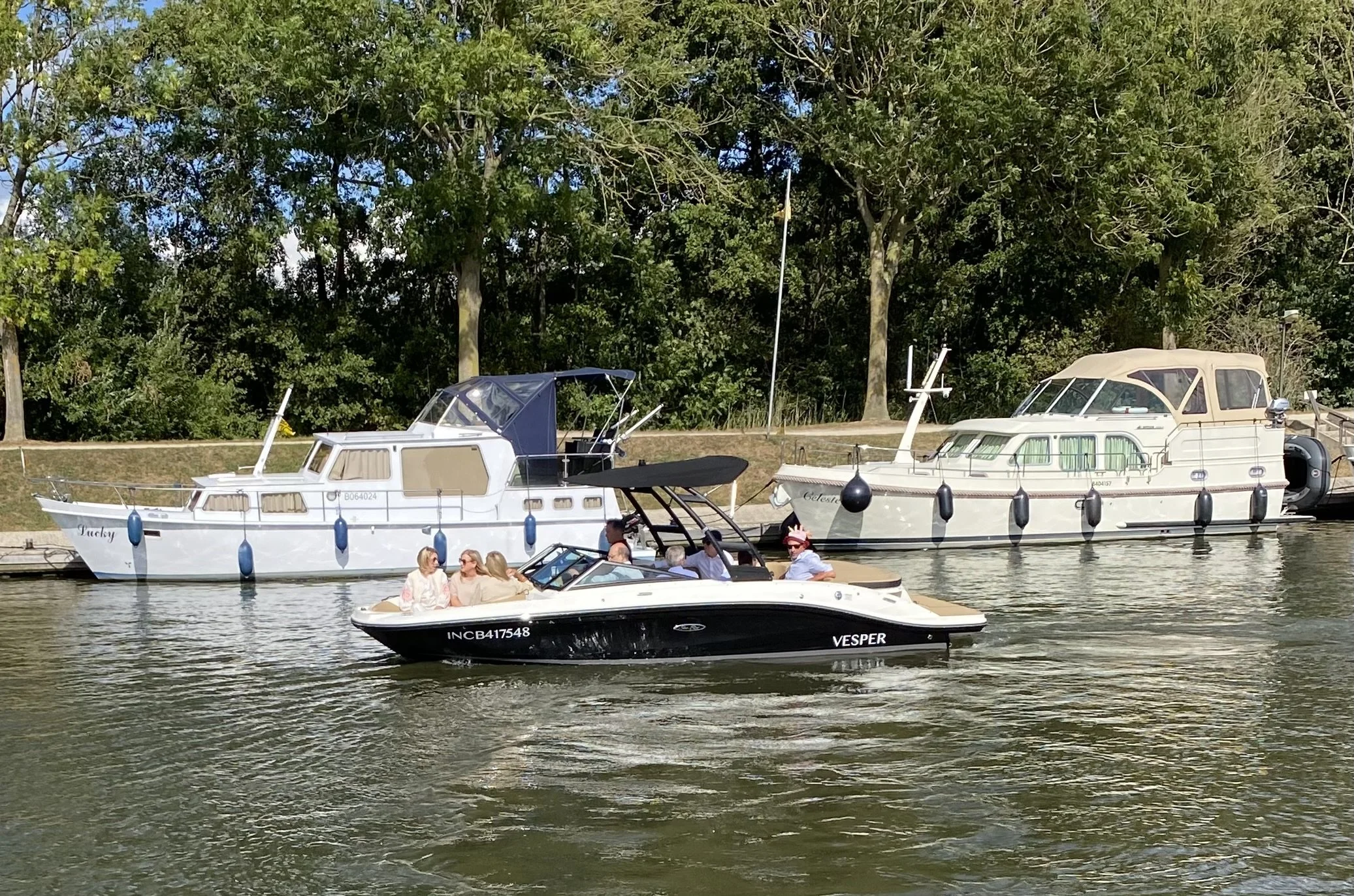 Meer met drie boten, twee grote boten aan de kant en een kleine speedboot met zeven mensen aan boord, op stromend water met bomen op de achtergrond.