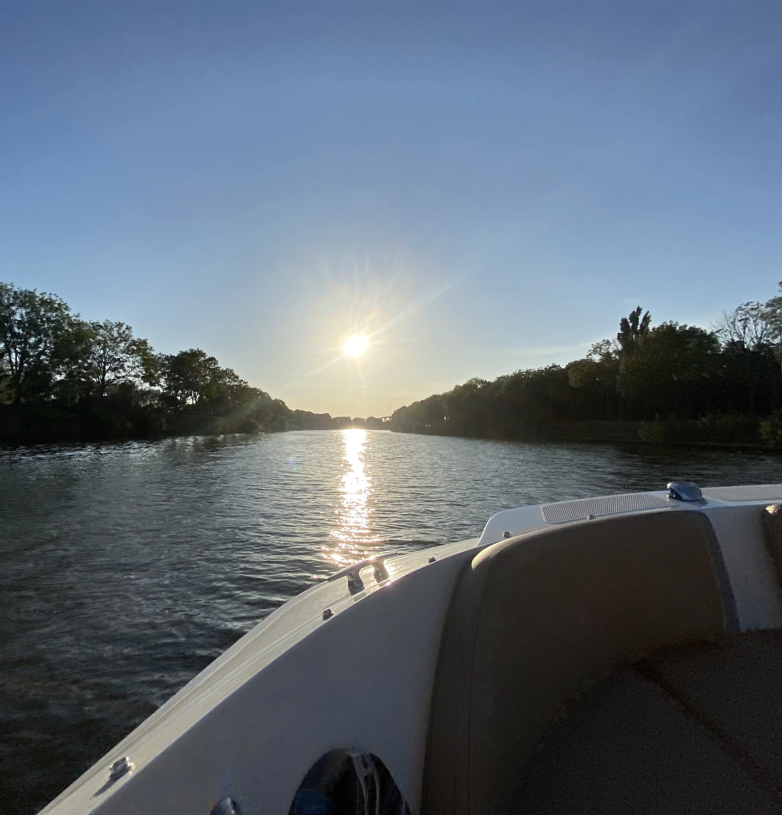 Een rivier gezien vanaf een boot, met de zon ondergaat en weerspiegeling in het water, omringd door bomen.