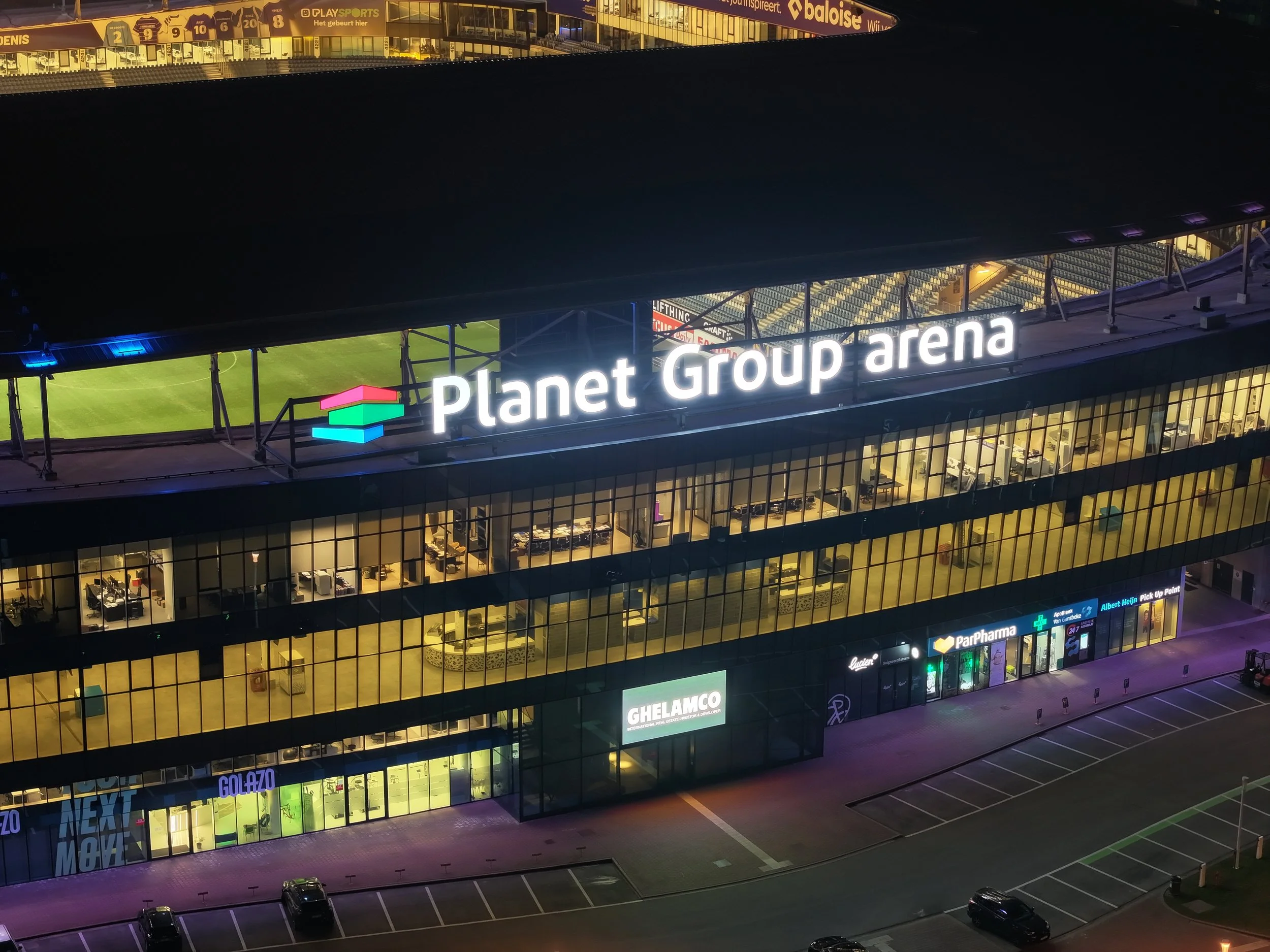 Uitzicht op het gebouw van Planet Group arena 's avonds, met verlichte gevel en kantoorruimtes zichtbaar binnen, parkeerplaatsen voor het gebouw.