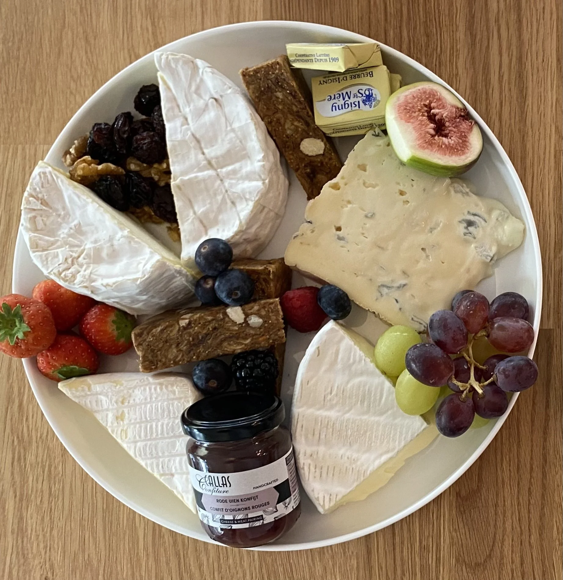 Een kaasplank met verschillende kazen, vers fruit zoals aardbeien, druiven, vijgen en bosbessen, en een potje confituur.