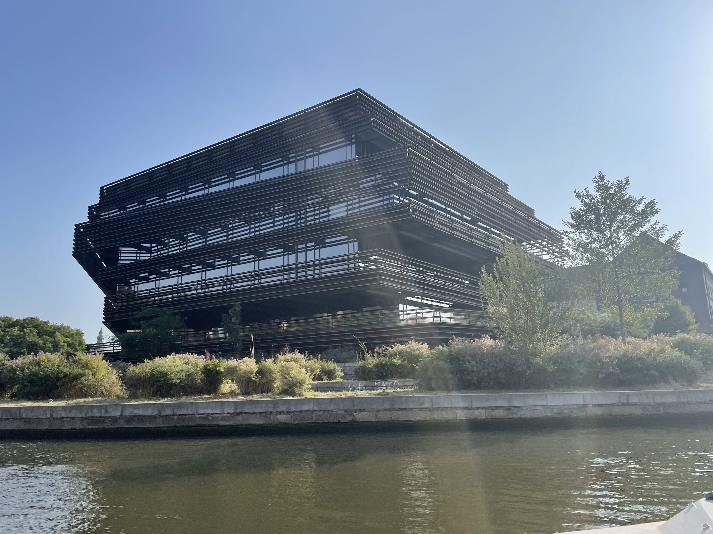 Moderne zwarte gebouw met horizontale lijnen naast water en groene bomen onder een heldere blauwe lucht.