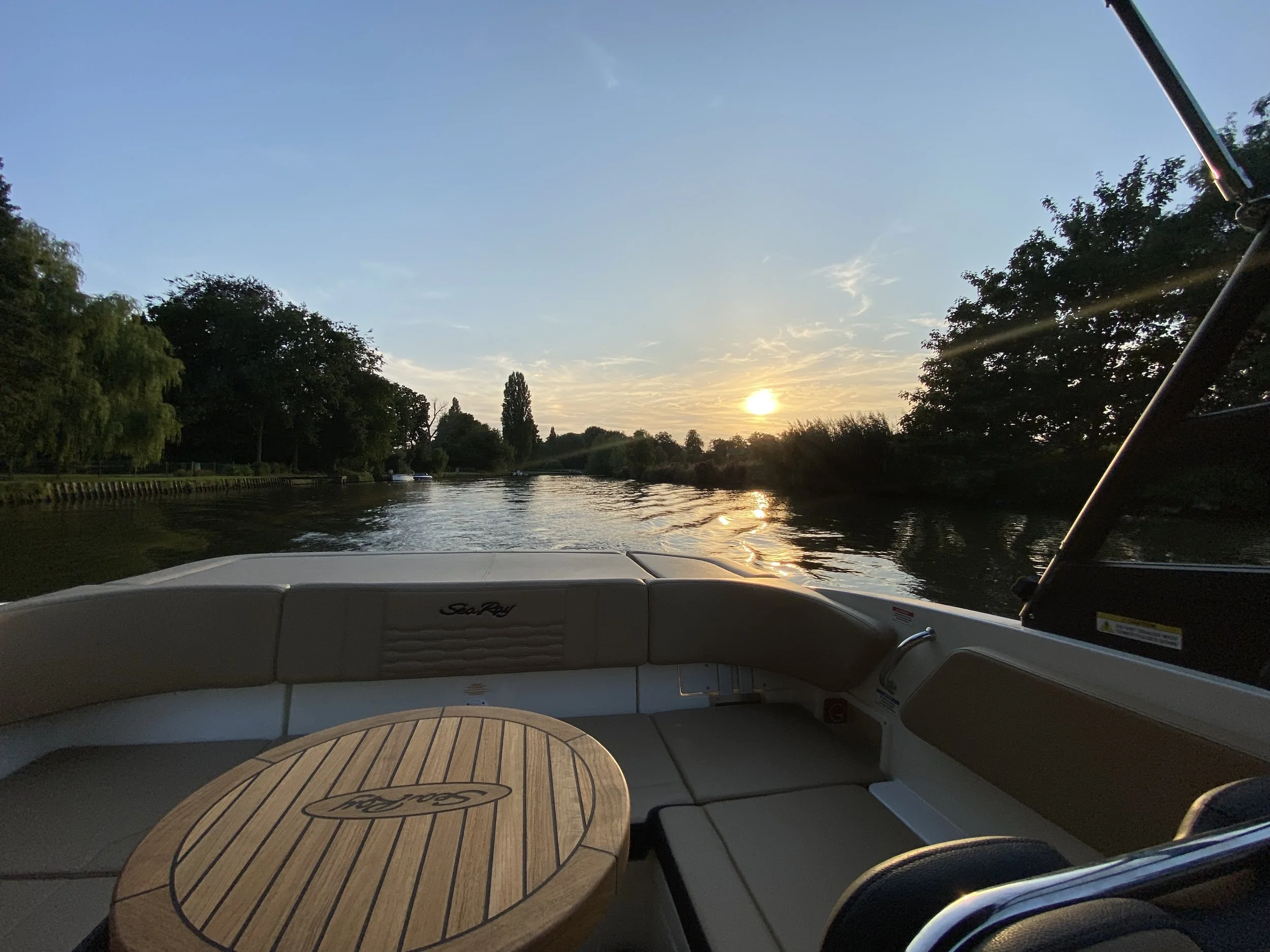 Uitzicht vanaf een boot op de rivier tijdens zonsondergang, met een houten tafel, comfortabele zitplaatsen en omringende bomen.