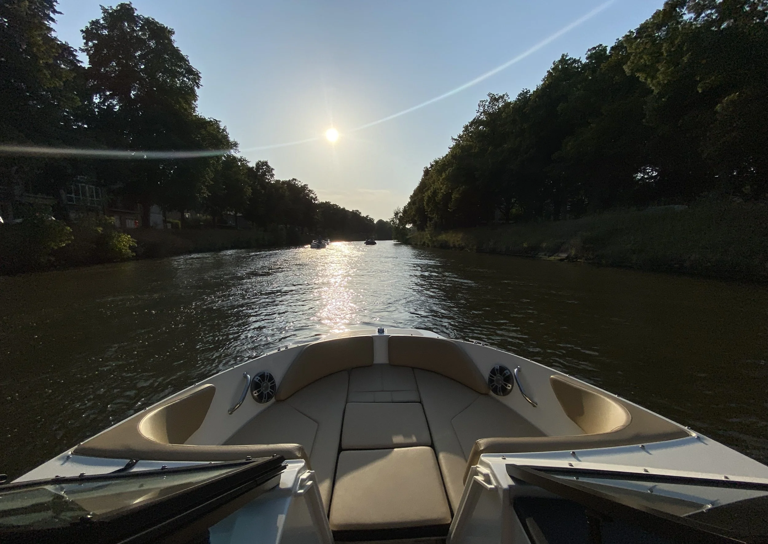 De foto toont de voorkant van een wit speedboot op een rivier, met de zon laag aan de hemel en groene bomen aan weerszijden.