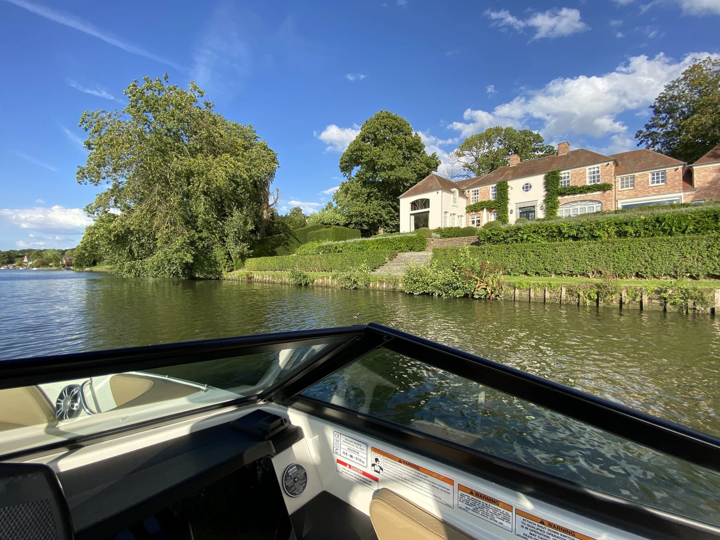 Uitzicht vanuit een boot op een rivier met aan de oever een groot huis met een tuin, bomen en een heldere blauwe lucht.