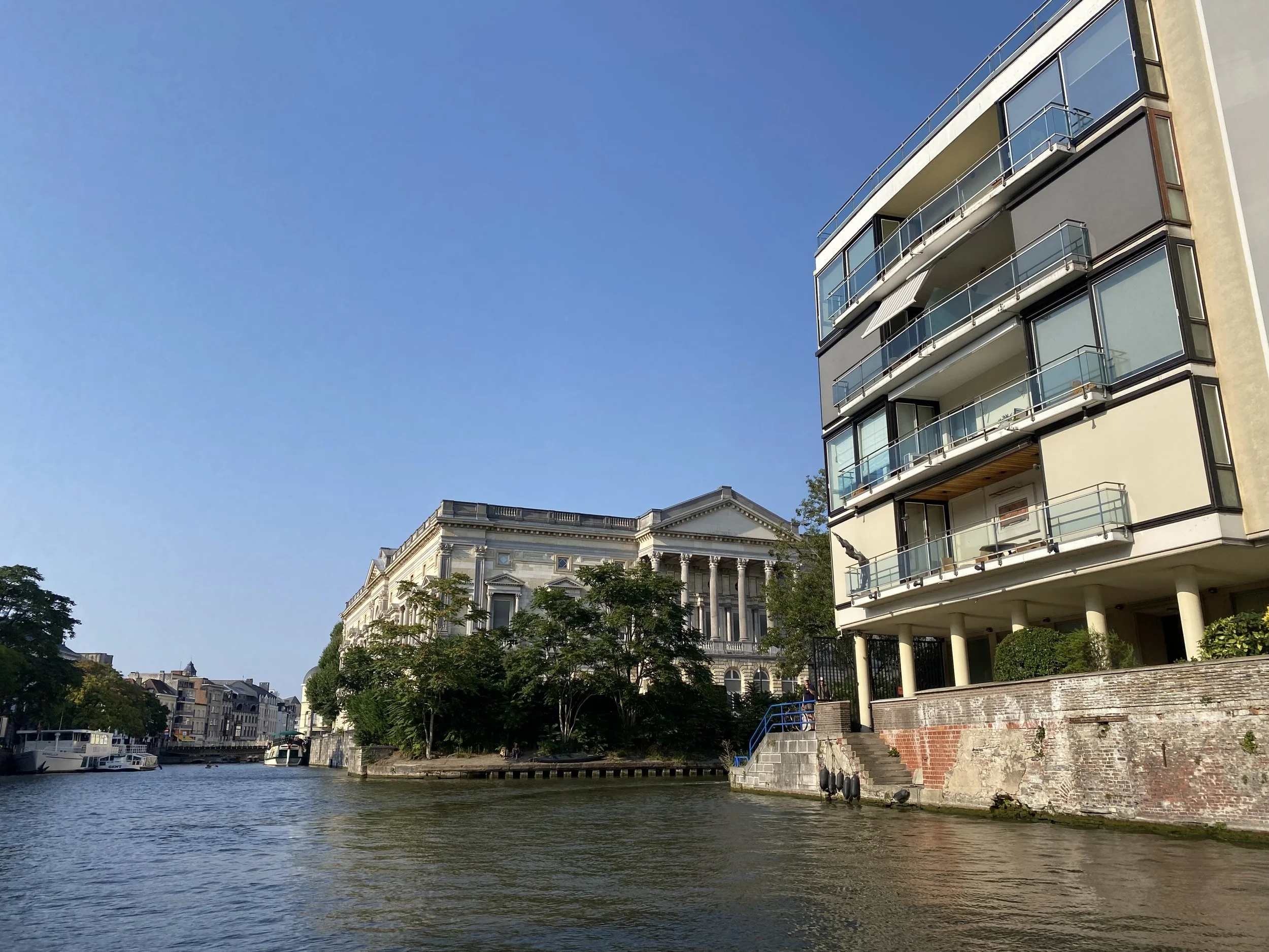 Uitzicht op een rivier met aan beide zijden gebouwen, waaronder een modern appartementengebouw en een oud gebouw met klassieke architectuur. Er zijn boten op de rivier en bomen langs de kade.