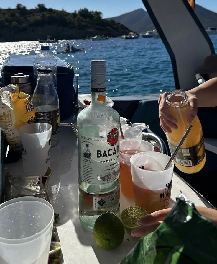 Buiten op een boot met uitzicht op het water en bergen, een tafel met drankjes, een fles Bacardi, limoen, ijs en personen die drinken en handen die flesjes vasthouden.