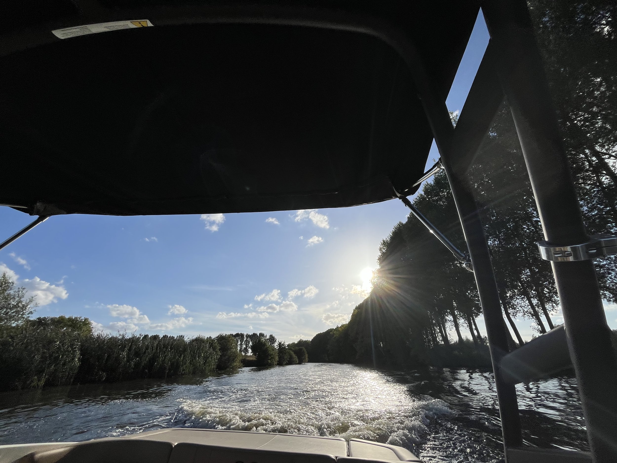 Uitzicht vanaf een boot op een rivier met veel groene bomen aan weerszijden en een blauwe lucht met enkele witte wolken, met de zon die achter de bomen schijnt.