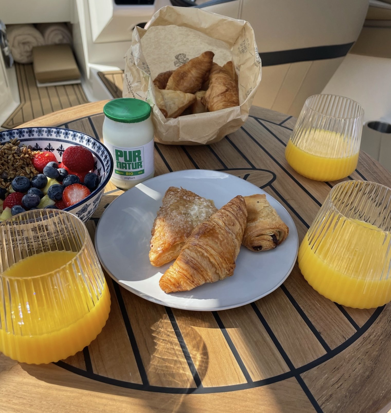 Een ontbijtbord met een croissant, een chocoladebroodje en een kaneelbroodje, een kom met fruit en muesli, twee glazen met sinaasappelsap, een bakje met yoghurt, en een papieren zak met meer croissants op een houten tafel.