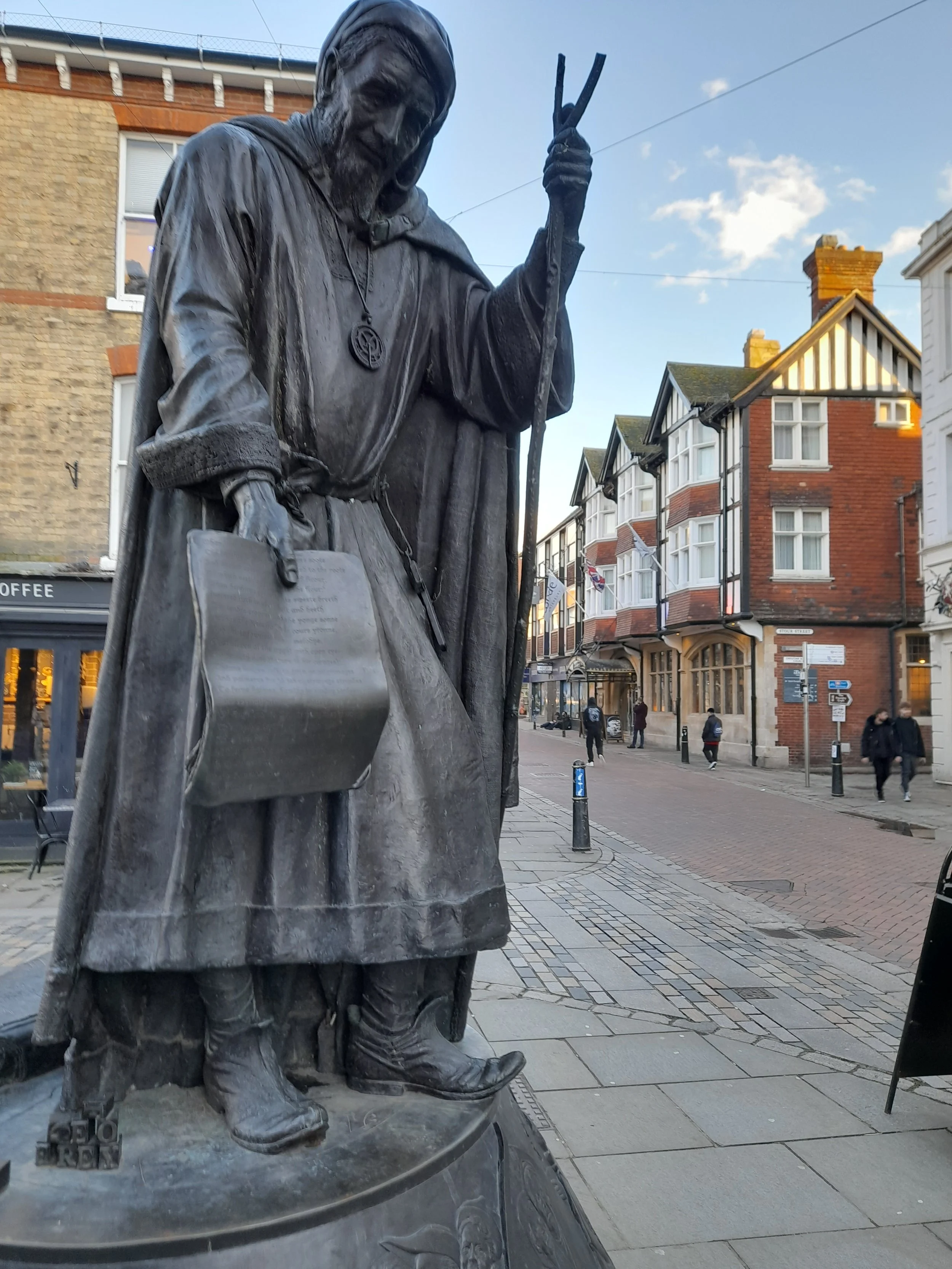Statue of a medieval pilgrim in Canterbury, wearing a cloak and hat, holding a walking staff, with historic buildings nearby.