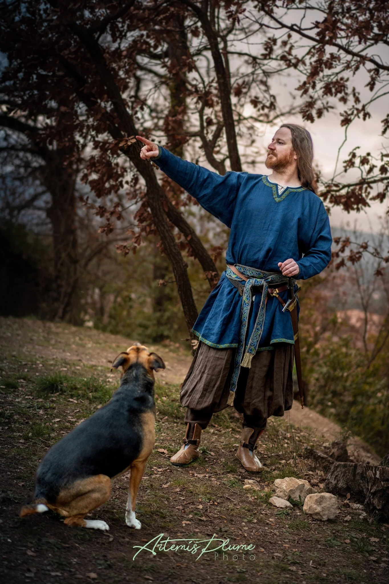 Photo d'un homme en costume médiéval pointant du doigt quelque-chose hors du champ, un chien est assis à ses pieds