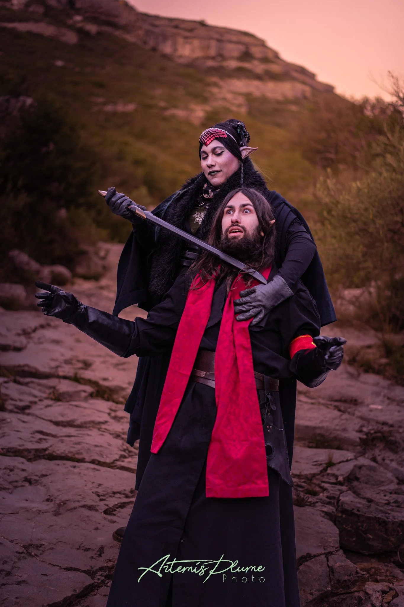 Photo d'une femme tenant un sabre factice noir au cou d'un homme. Les deux sont en costume en costume d'elfe sombre