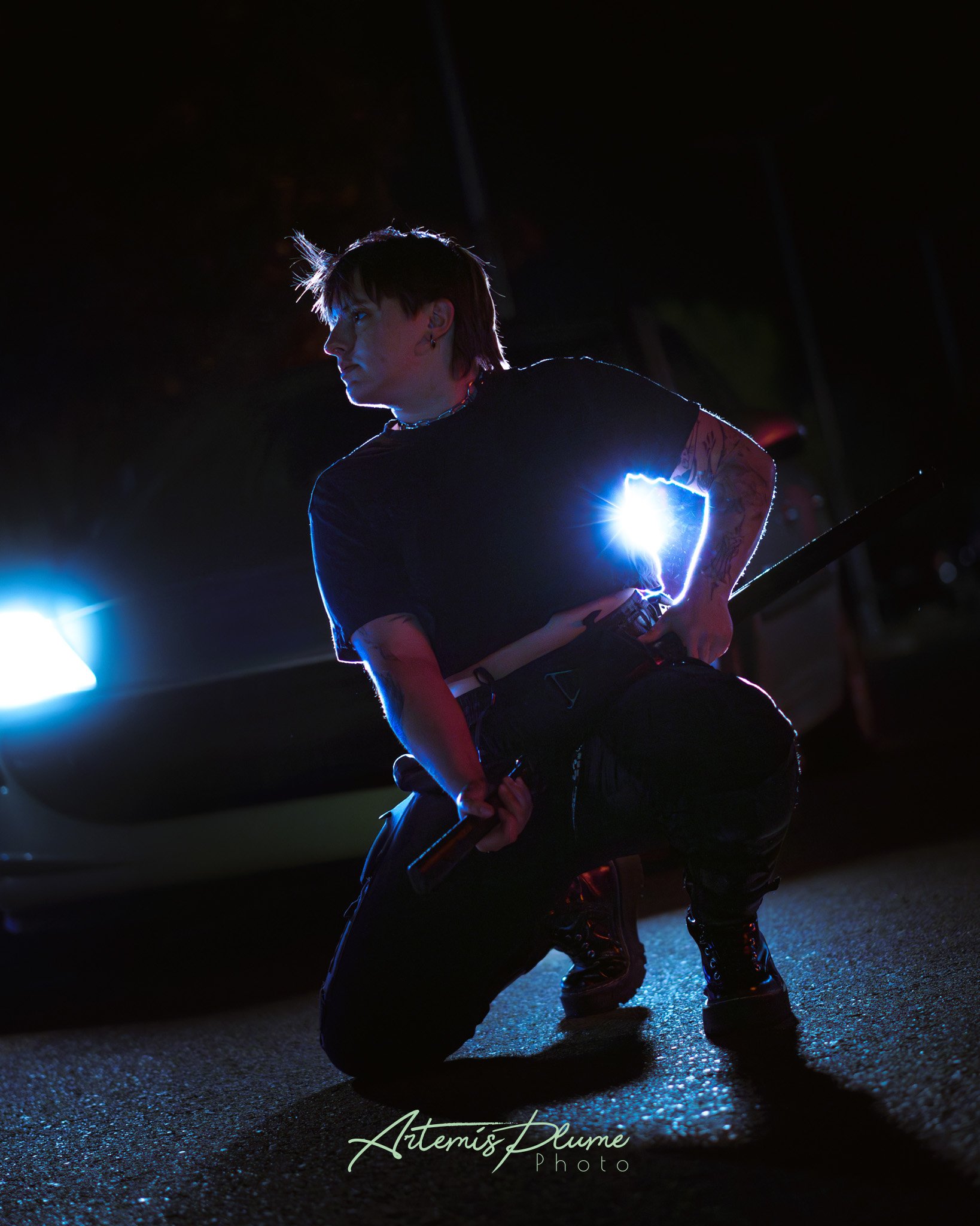 Photo d'une femme habillée en noir, accroupie devant une voiture phare allumées. Elle prépare un sabre