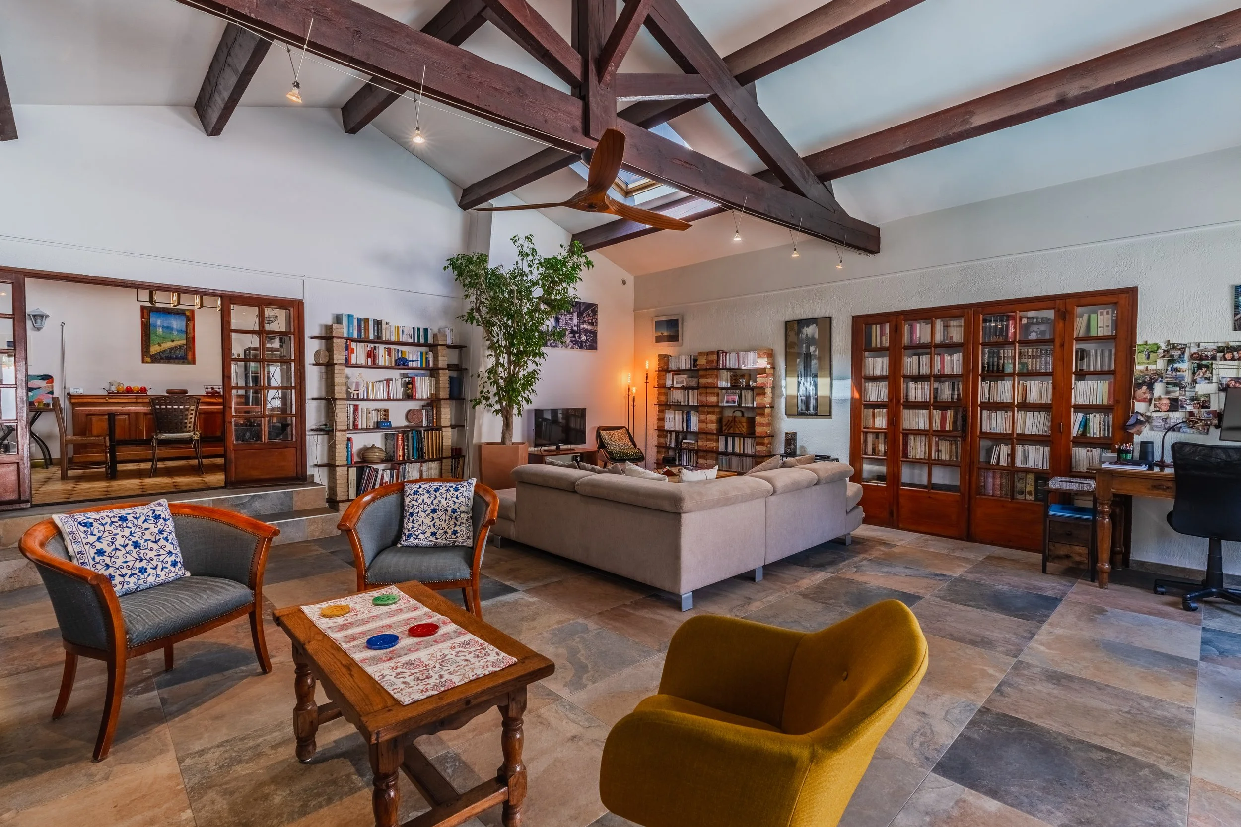 Salon intérieur avec un plafond en bois, une grande bibliothèque, un canapé beige, un fauteuil jaune et un tapis en pierre, décoré avec des plantes et des œuvres d'art.