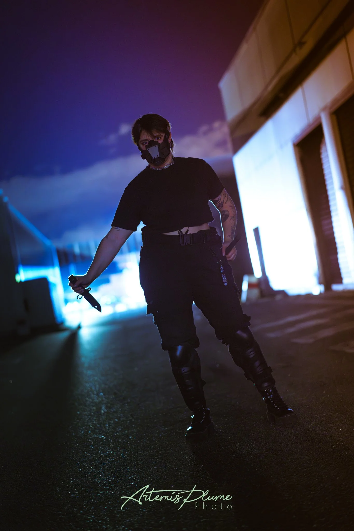 Photo d'une femme habillée en noir et portant un masque dans une ruelle sombre. Elle a un couteau dans sa main