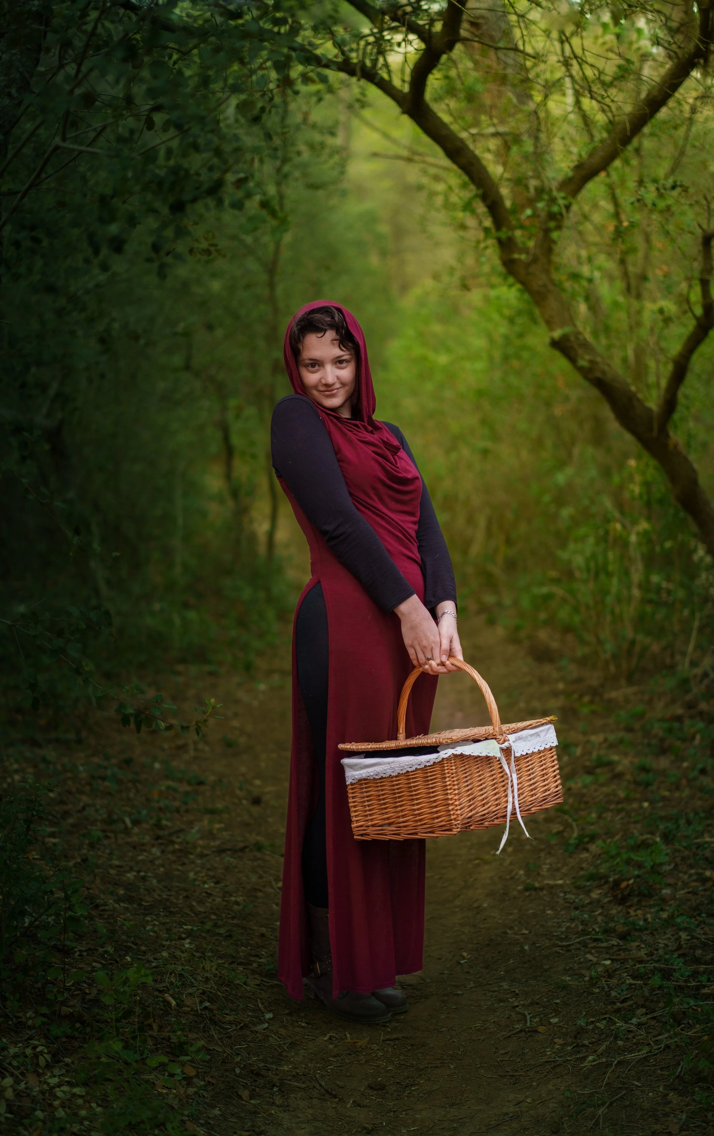 Une femme marchant dans une forêt, portant un panier en osier et vêtue d'une robe bordeaux avec un châle noir, entourée de verdure.