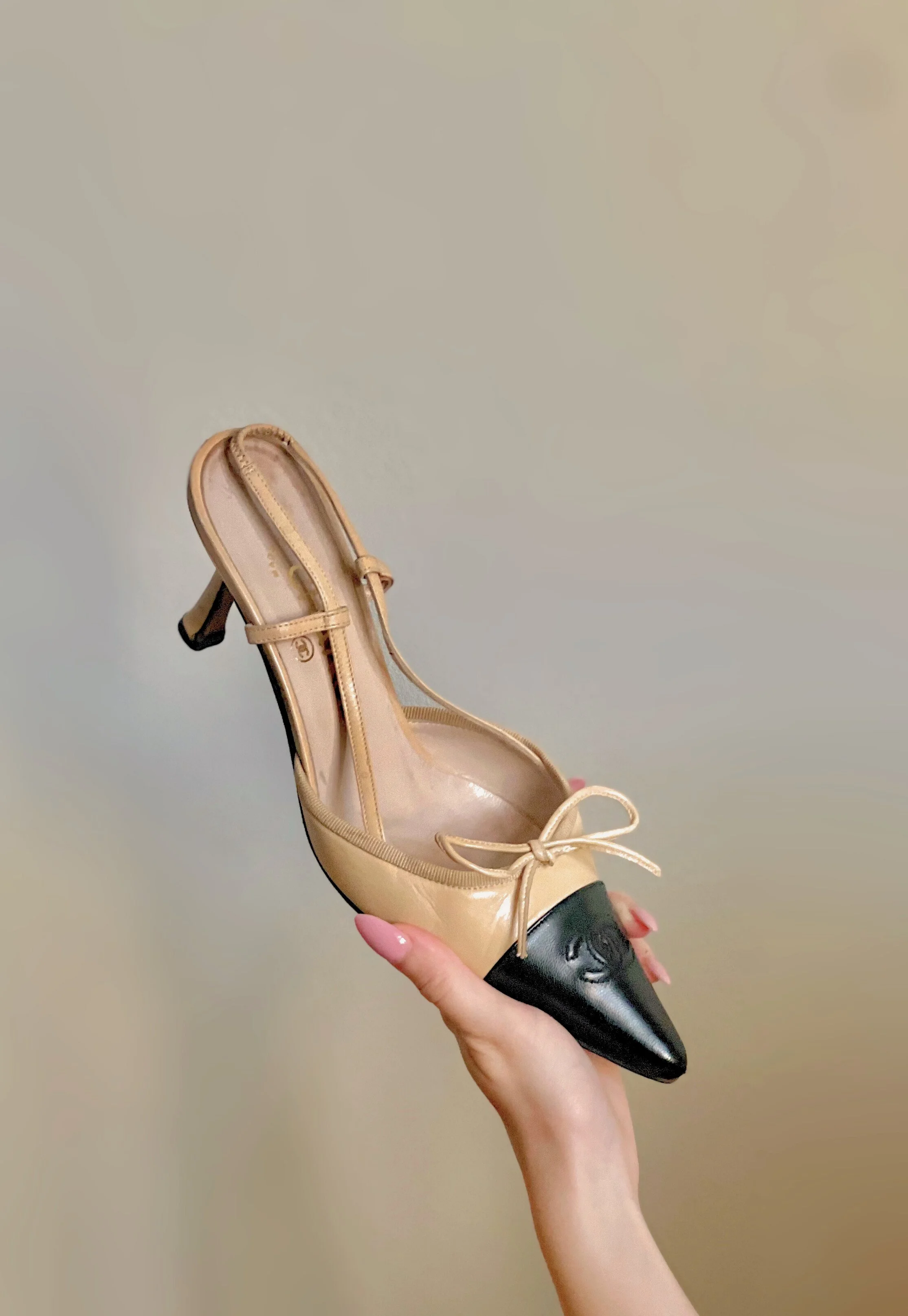 A high heel shoe with a beige body and a black pointed toe, held in a person's hand against a plain wall background.