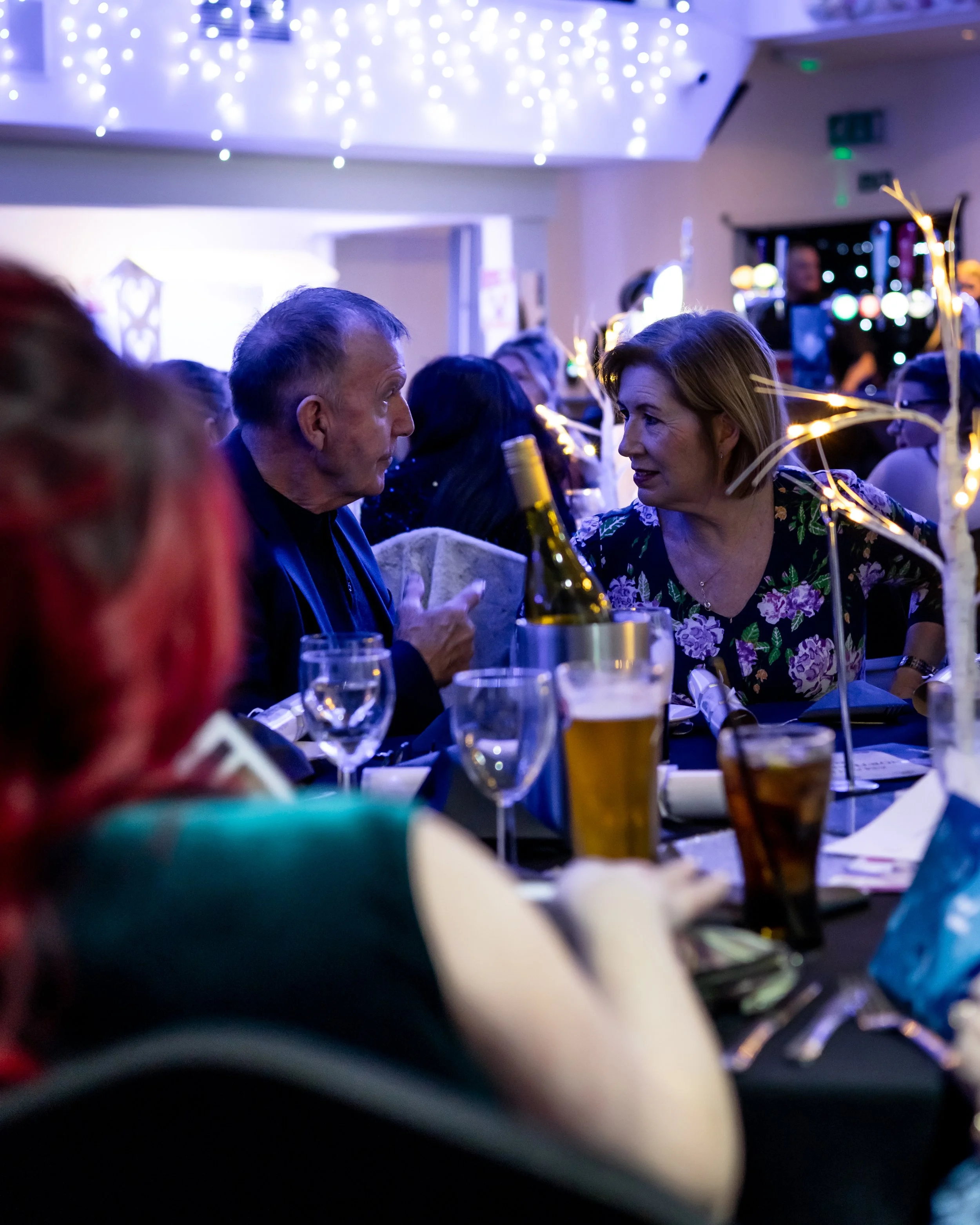 People sitting at a table during a festive event with string lights overhead, drinks, and table decorations, engaged in conversation.
