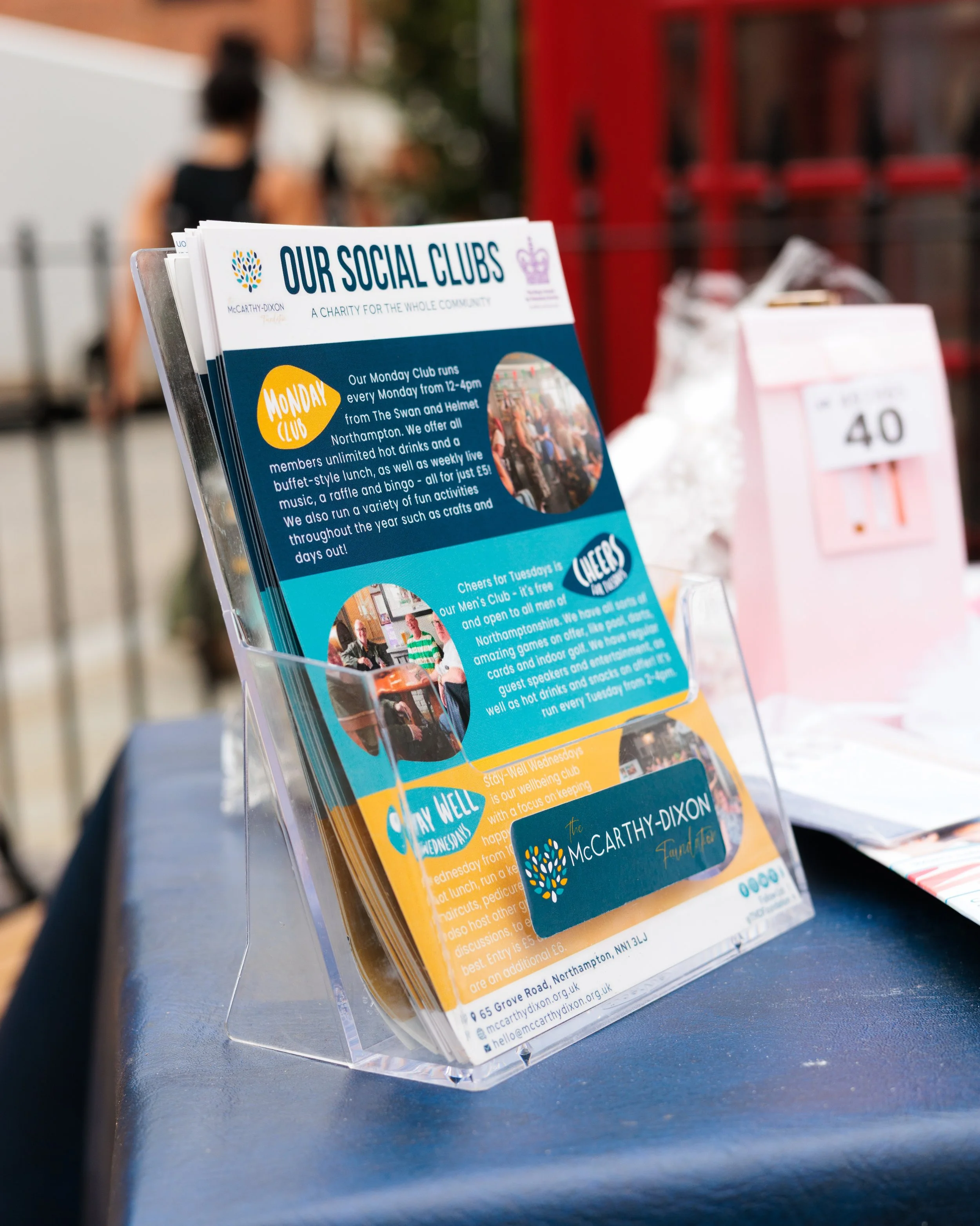 A transparent brochure holder displaying pamphlets about social clubs, placed on a table outdoors with a person walking in the background.