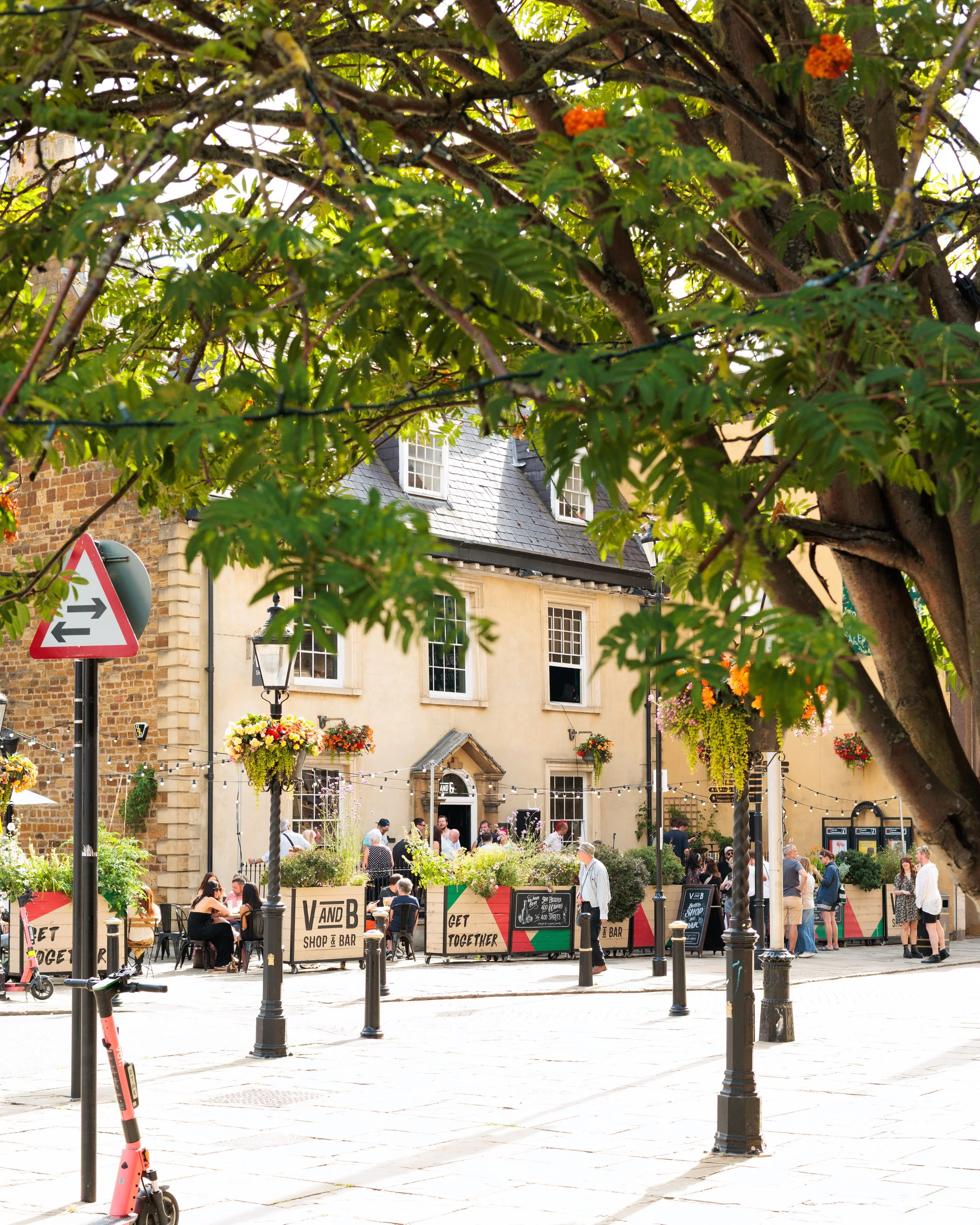 A lively outdoor cafe scene in a small town, with people sitting and standing outside a bar or restaurant, decorated with hanging flower pots, string lights, and a sign that reads 'V and B Shop & Bar'. The scene is framed by the branches and leaves o