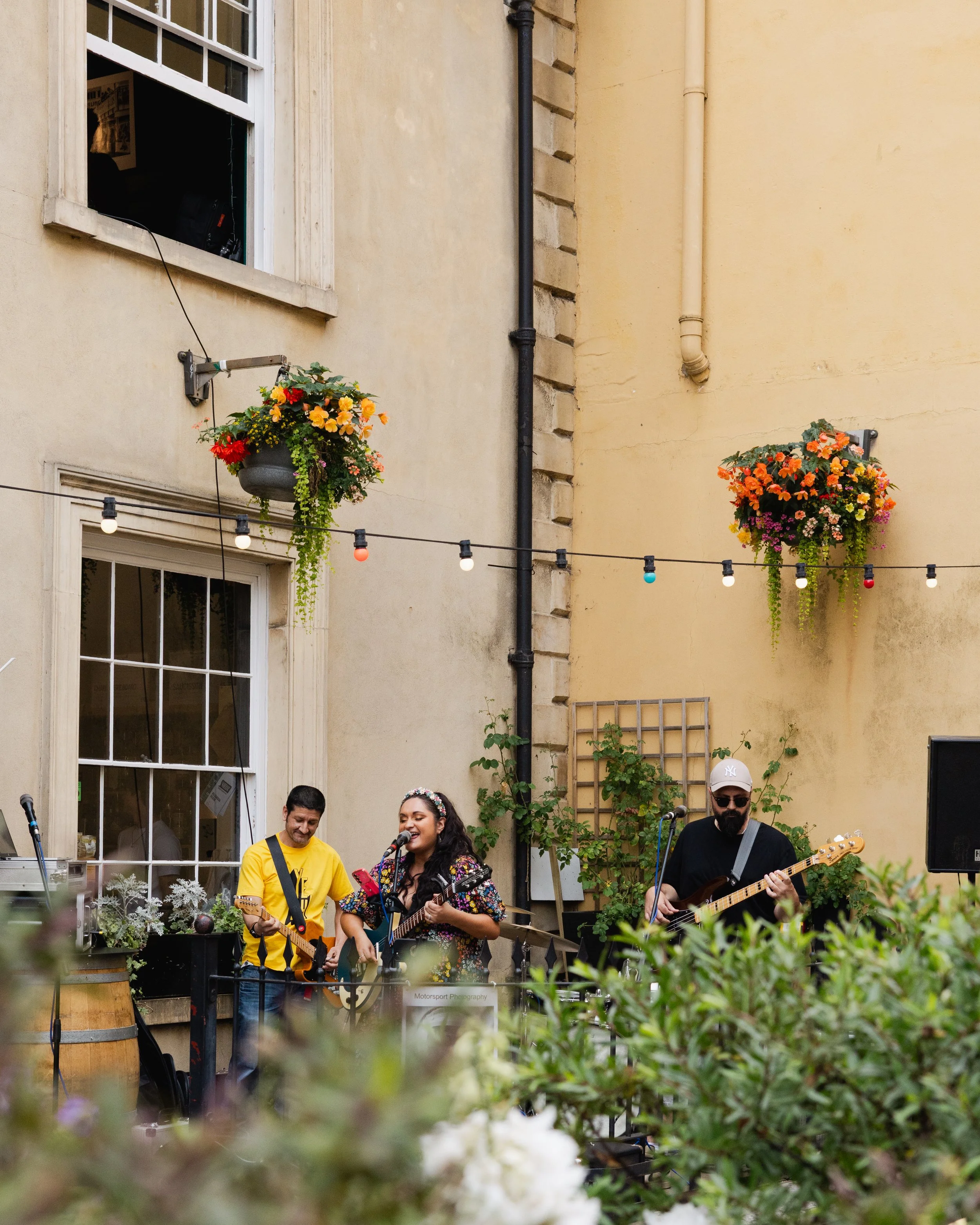Three musicians performing outdoors in a courtyard surrounded by yellow walls with hanging flower baskets and string lights. Two wear black shirts, one in yellow, all playing guitars and singing.