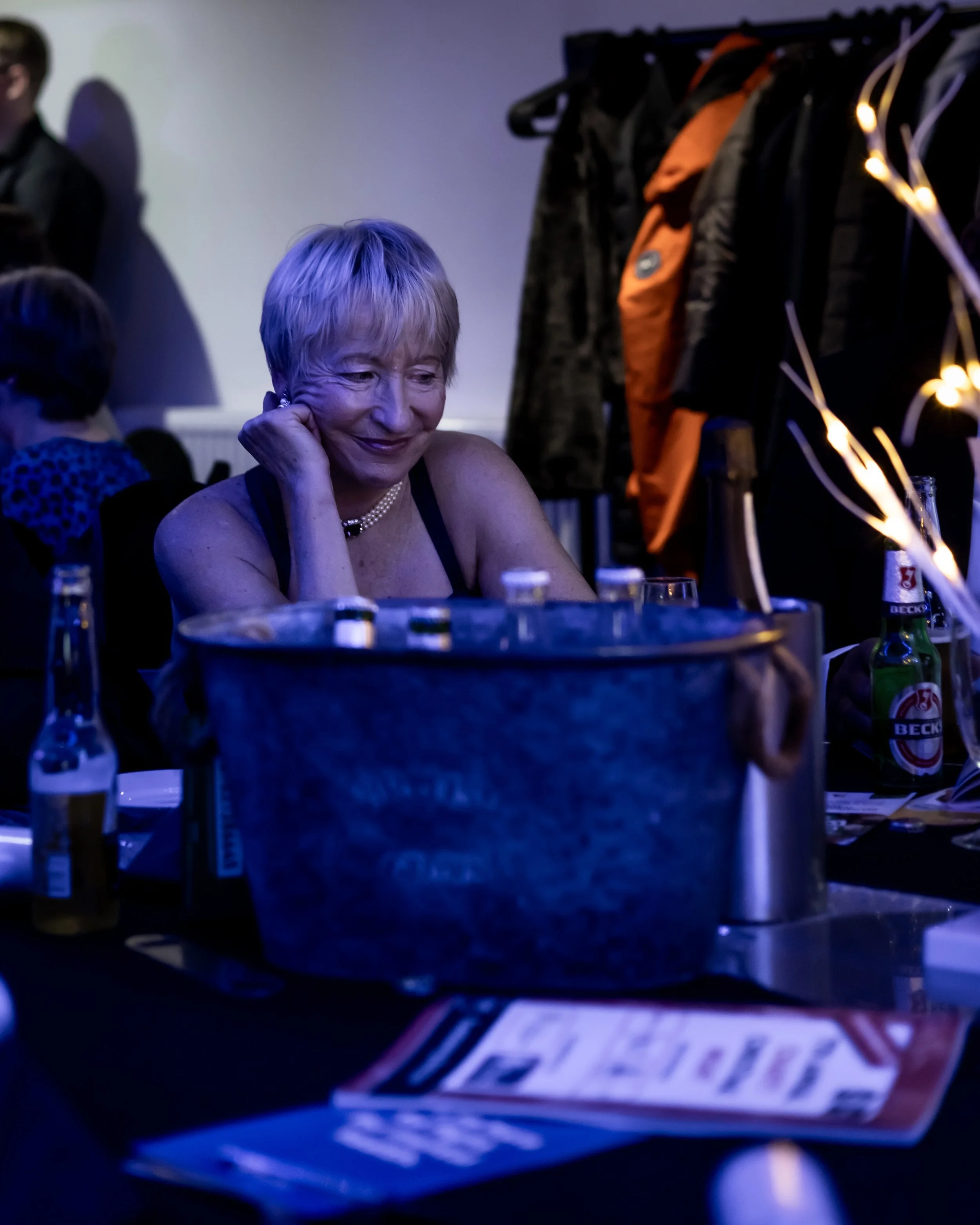 A smiling woman with short blonde hair sitting at a table during a party or celebration, with empty bottles and a metal bucket in front of her, and people and jackets in the background.