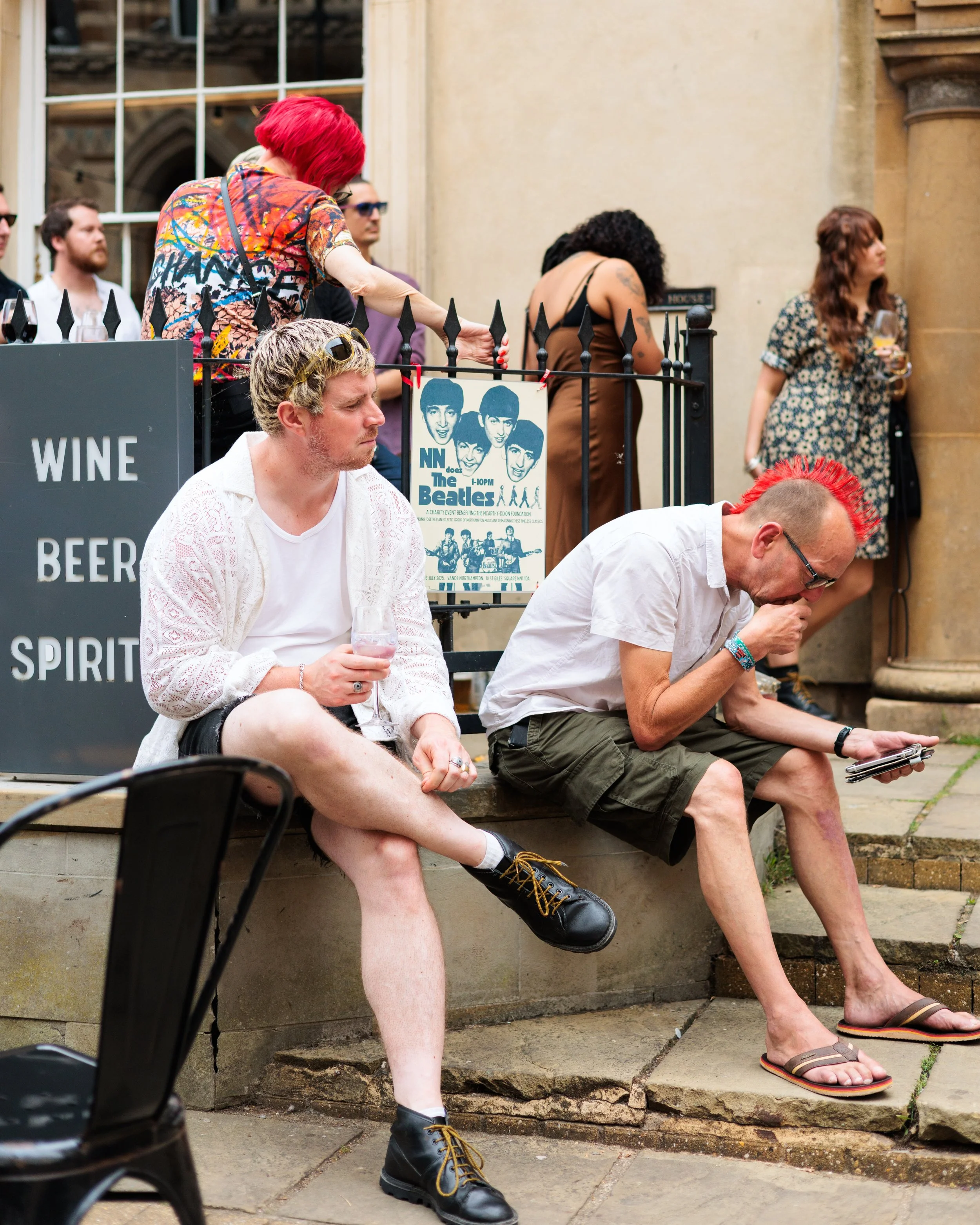 Two men sitting outside on a stone ledge, one with dyed red hair and a mohawk wearing glasses, a white shirt, and shorts, looking at a smartphone, the other with blond hair, sunglasses, a white shirt, and shorts, holding a drink. Behind them, a woman