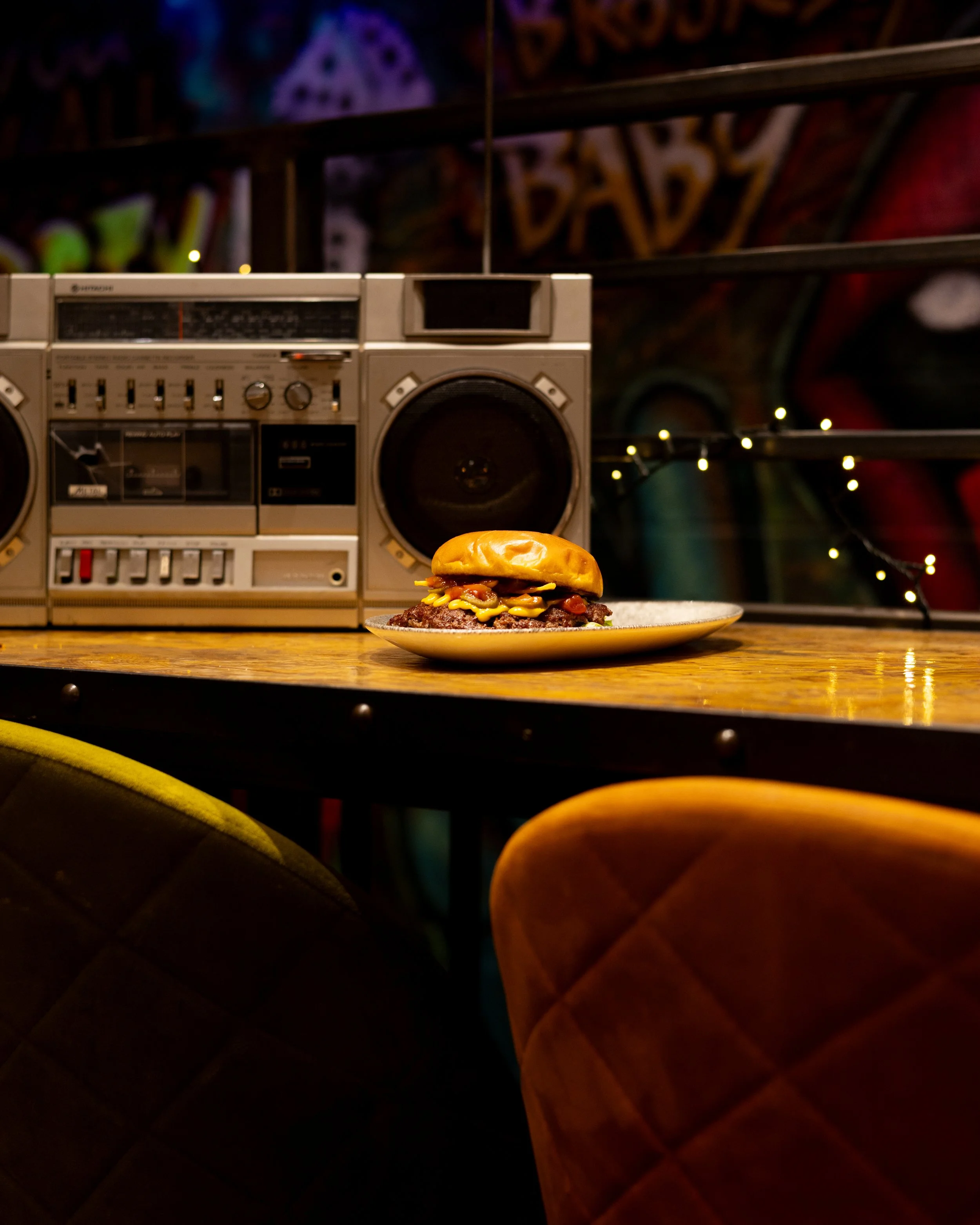 A plate with a cheeseburger on a wooden table in front of a retro stereo system, with a colorful graffiti wall in the background.