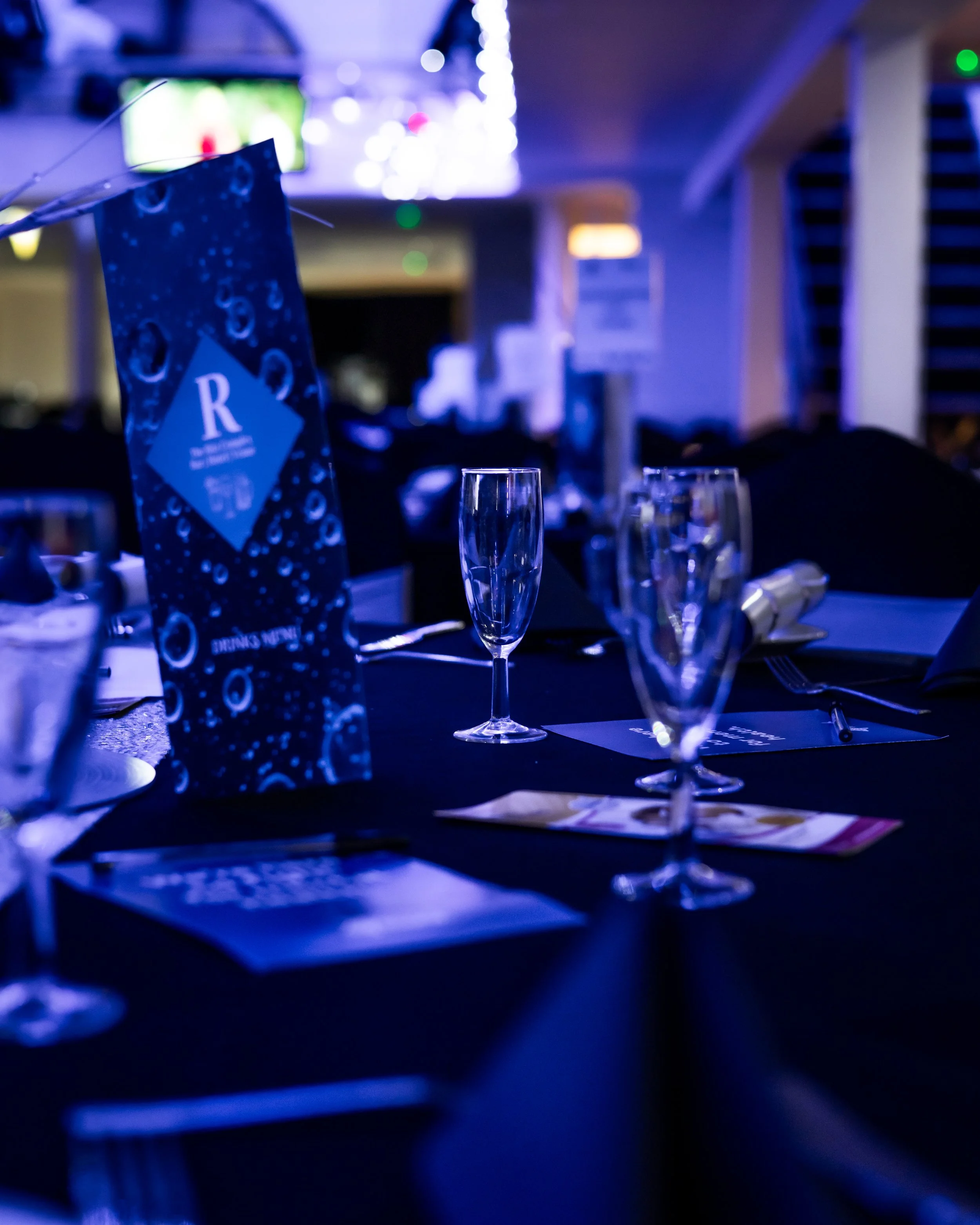 A dimly lit event space with a decorated table set with champagne glasses, a blue event program with bubbles, and a dark tablecloth.