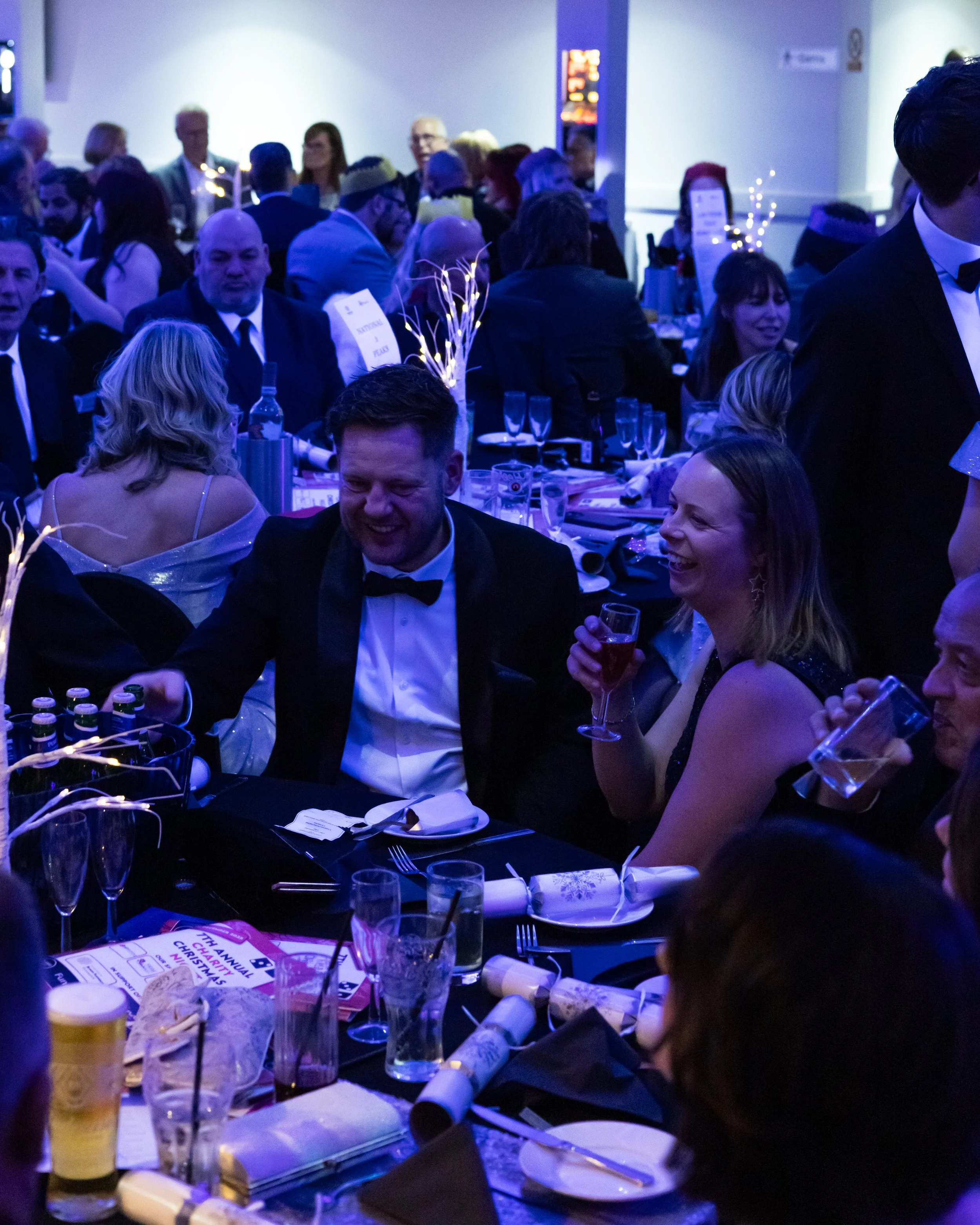 People dressed in formal attire, sitting at a decorated banquet table, laughing and enjoying a celebration or gala event in a dimly lit room with blue lighting.