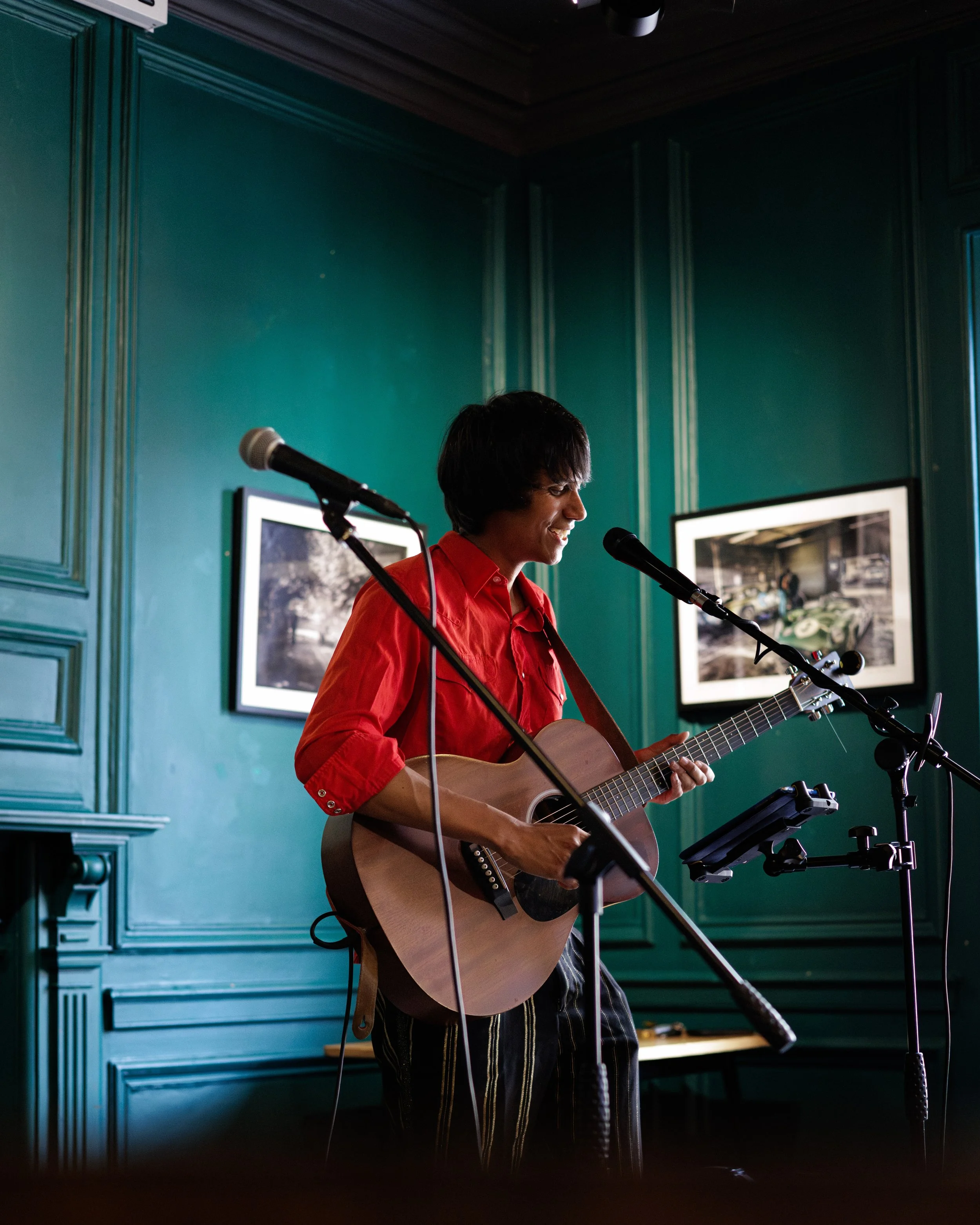 A person with short dark hair wearing a red shirt playing acoustic guitar and singing into a microphone in a room with teal walls decorated with framed photographs.
