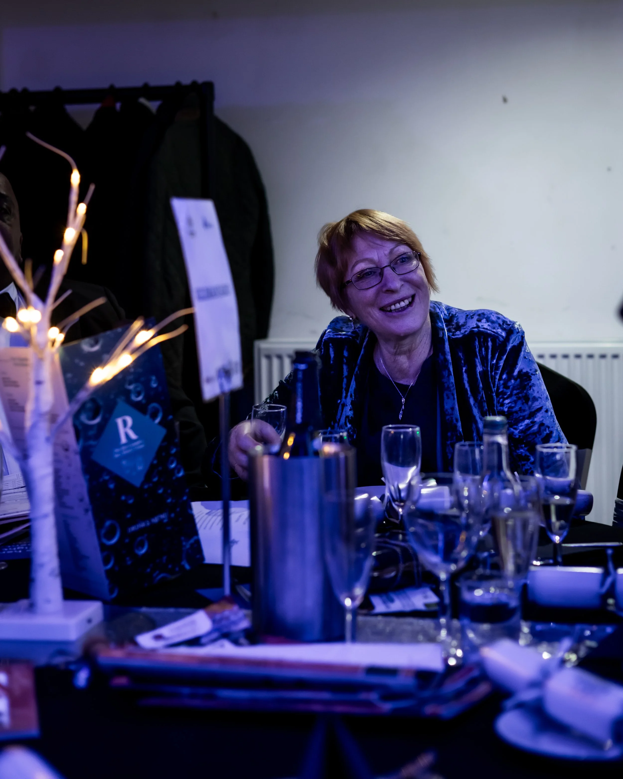 Woman with short hair and glasses smiling at a table set for a celebration or event, holding a glass of wine, surrounded by bottles, glasses, and event programs.