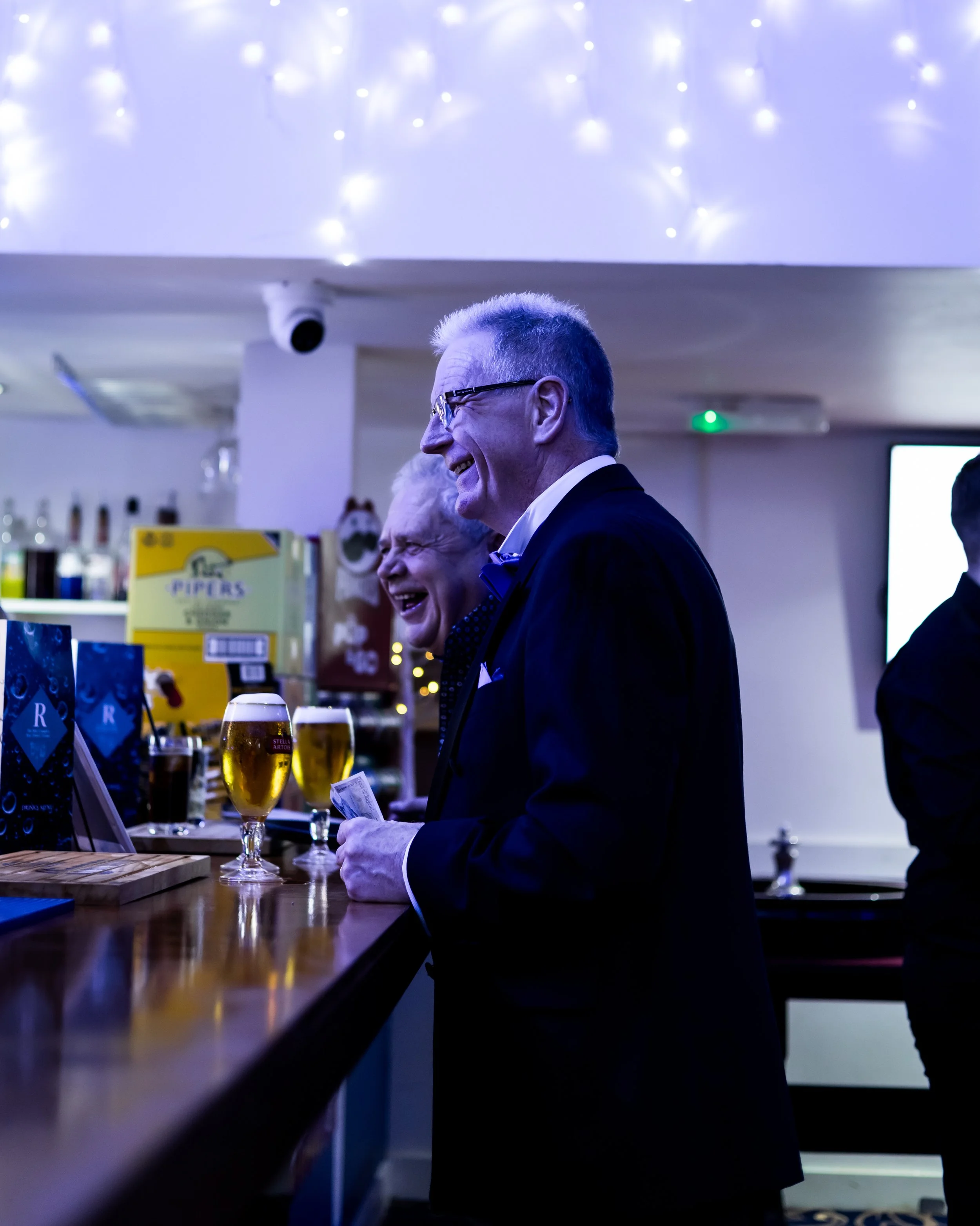 Two older men in tuxedos smiling and laughing at a bar, with drinks and bar signs behind them, under festive string lights.