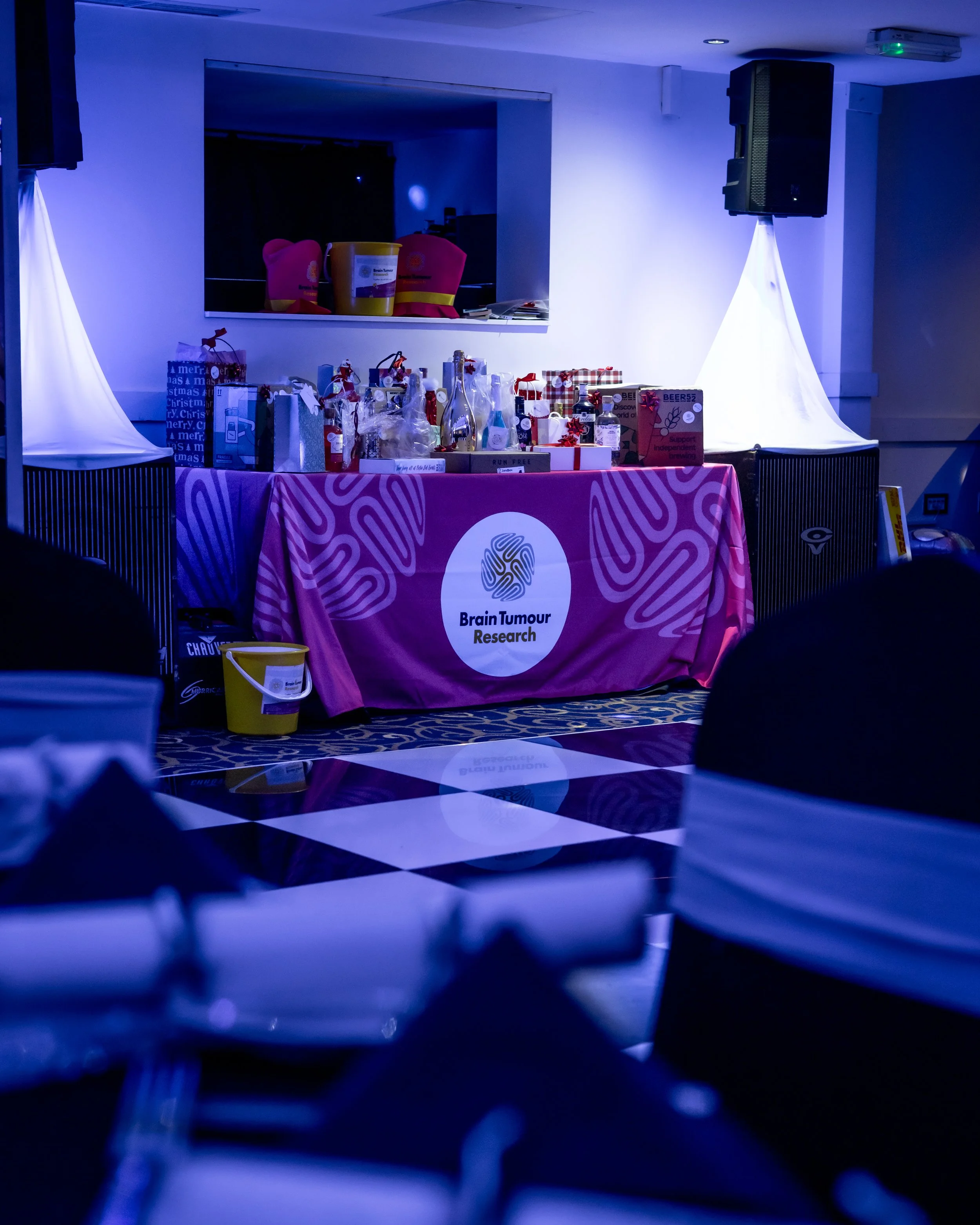 Table with a pink tablecloth displaying gifts and research materials for Brain Tumour Research at an indoor event, with a checkered black and white floor and blue lighting.