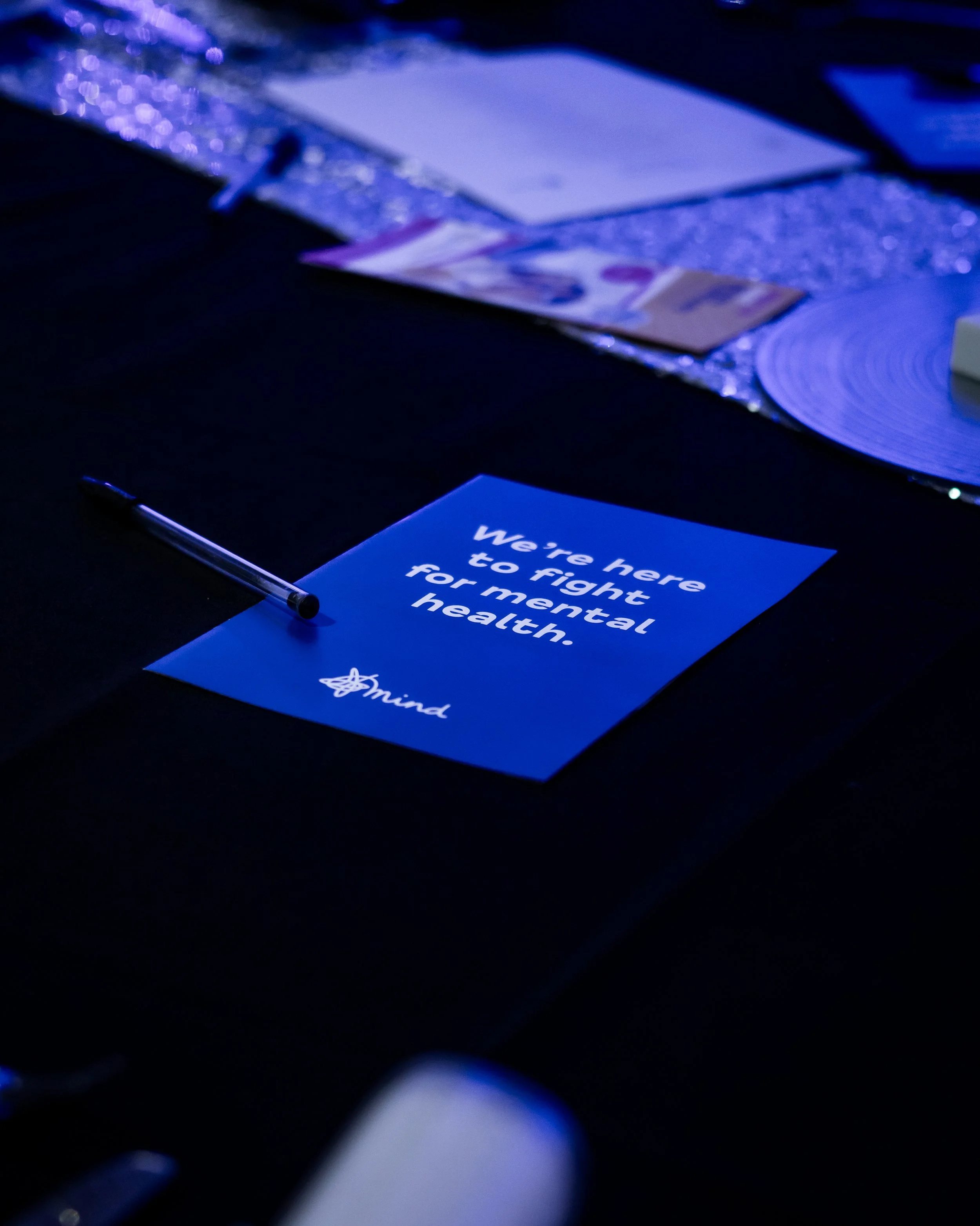 A blue pamphlet on a black table with the text 'We're here to fight for mental health'. A pen rests next to it, and there are various papers and materials on the table.