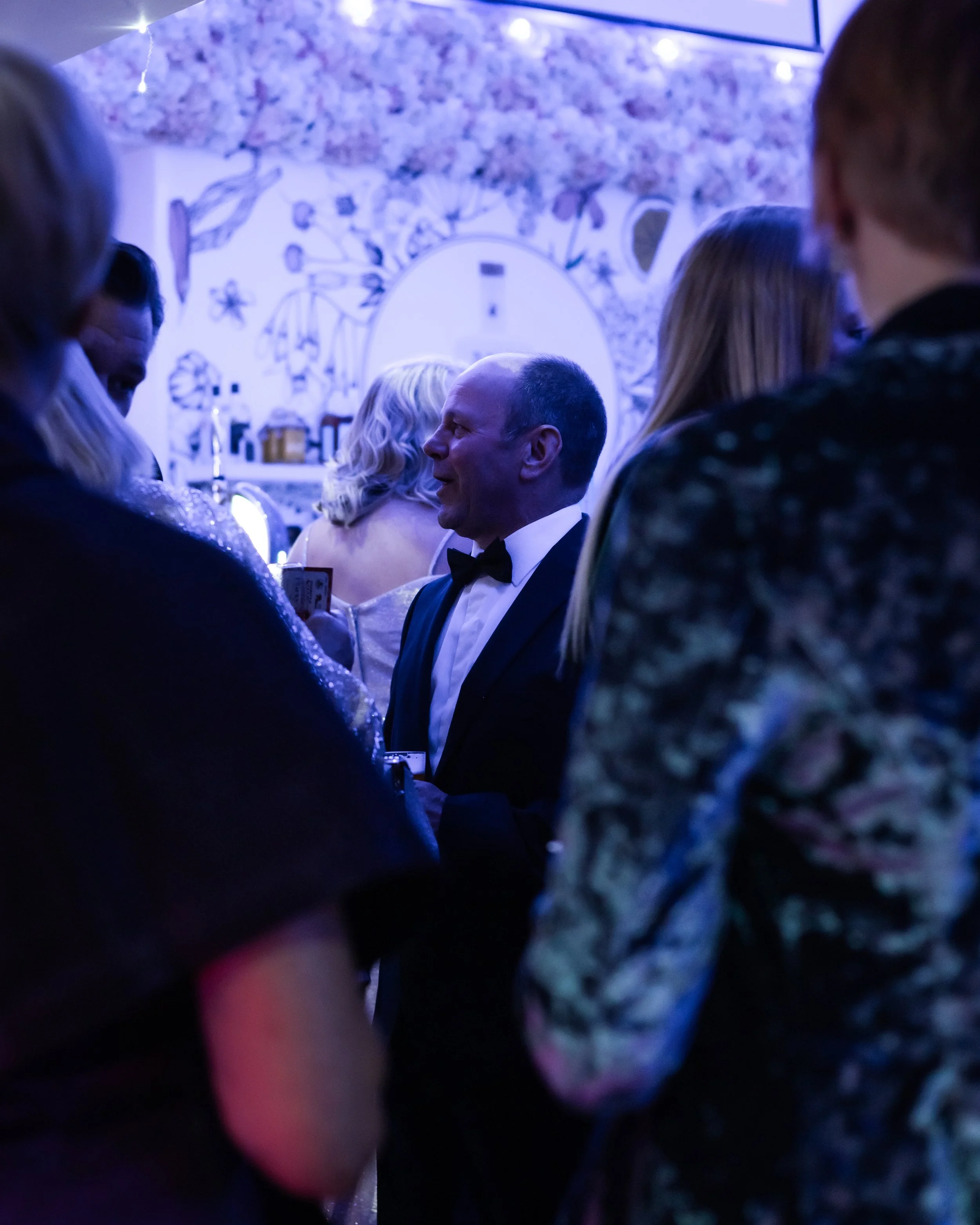A group of people in formal attire socializing at a party with blue lighting and floral wall decor.