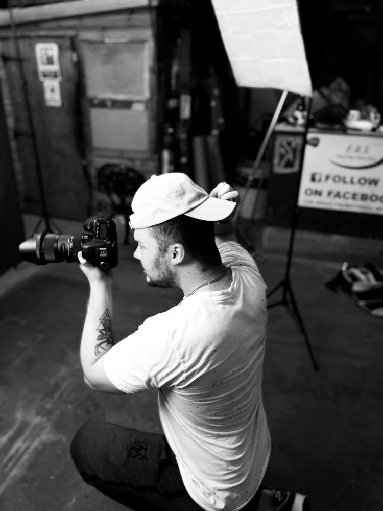 A young man wearing a baseball cap is kneeling and looking through the viewfinder of a professional camera in a photography studio set.