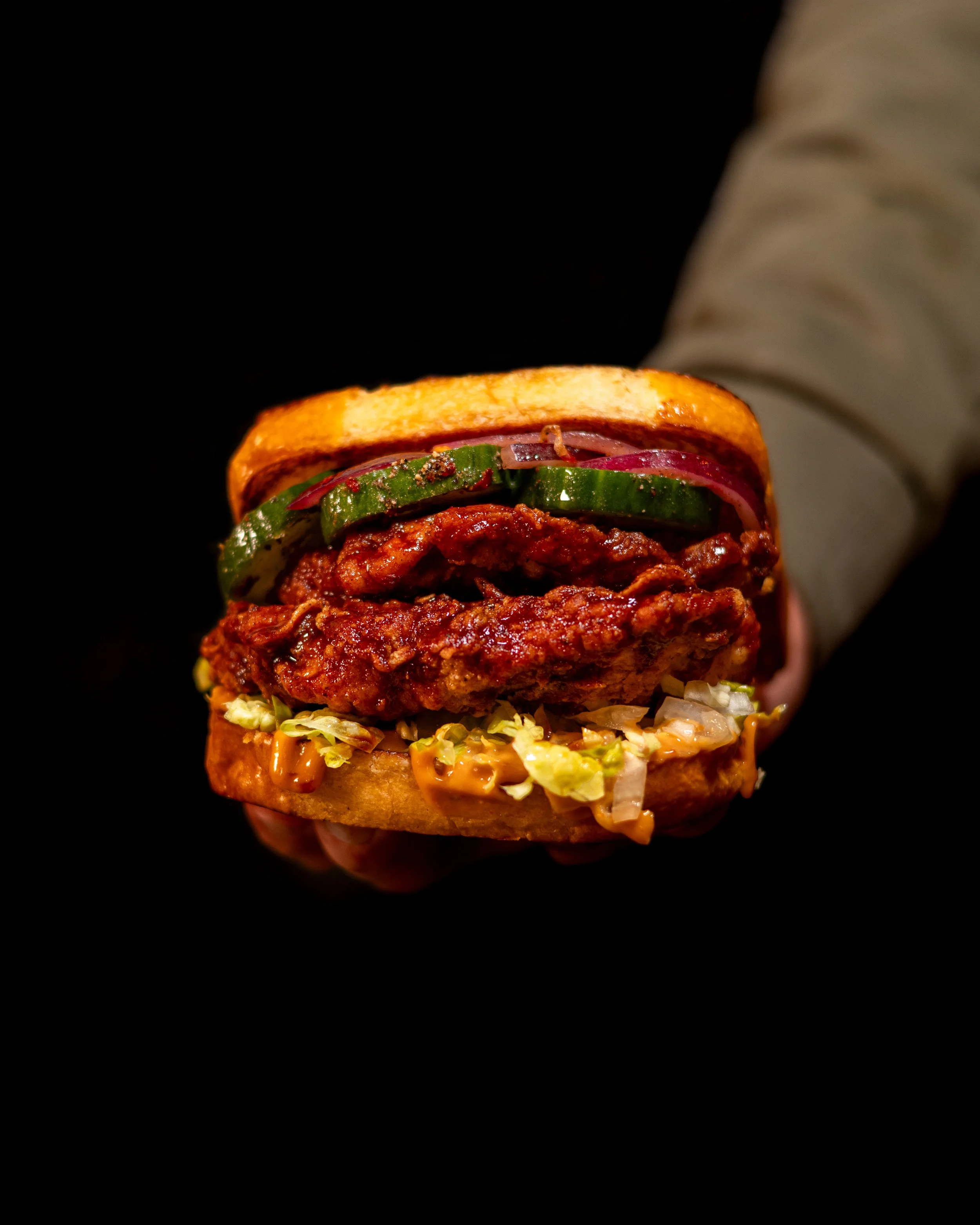 A person holding a fully assembled fried chicken sandwich with lettuce, tomato, pickles, onions, sauce, and a toasted bun against a black background.