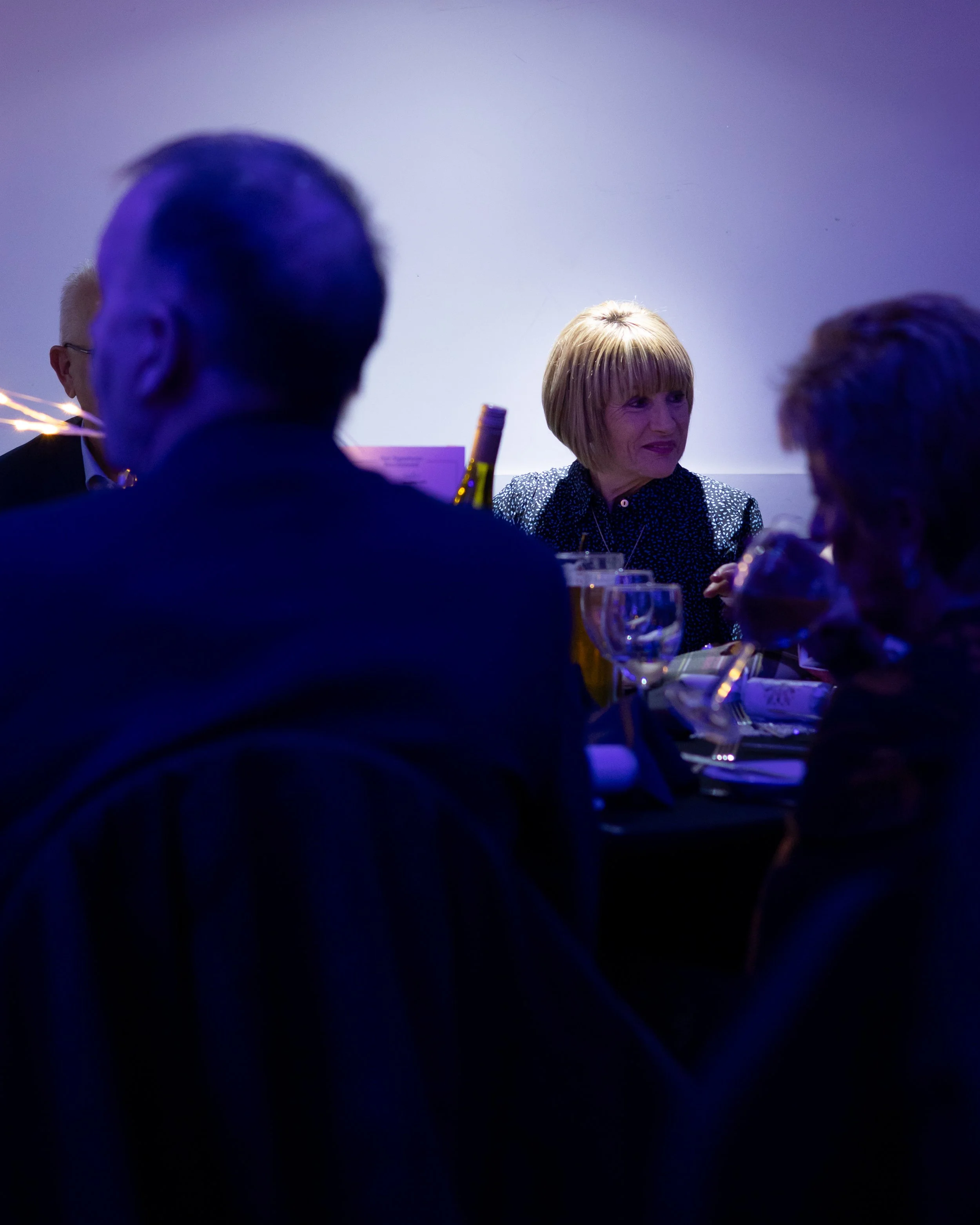 People sitting around a table at a formal event, with a woman in a patterned top in focus, illuminated by blue-purple lighting.