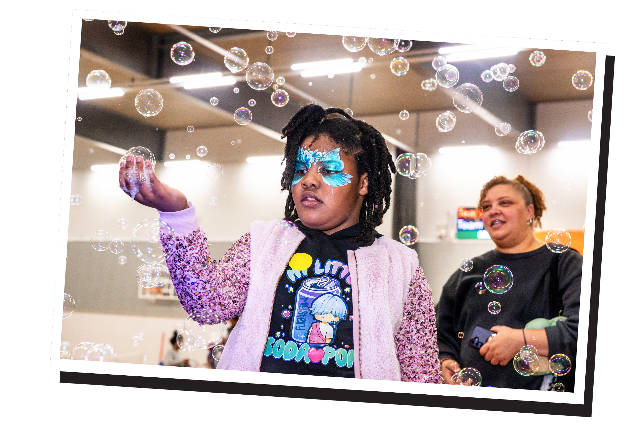 A young woman with blue face paint and a pink jacket playing with soap bubbles at an indoor event, with another woman in the background watching.