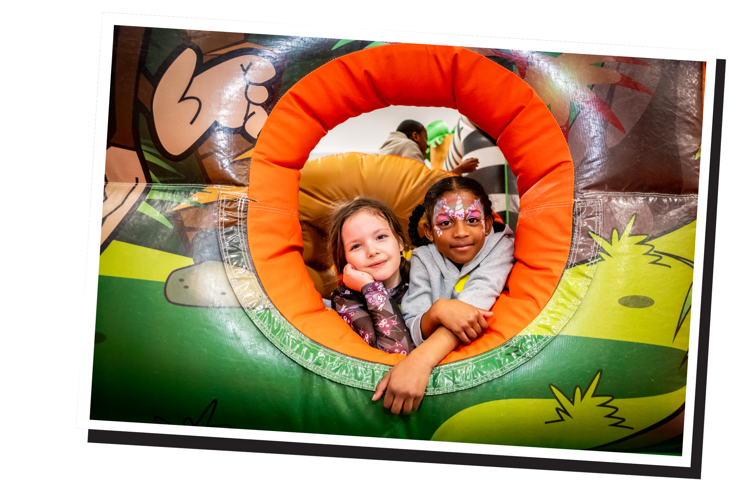 Two children sit inside a colorful jungle-themed inflatable play structure, with one girl wearing face paint and smiling and another girl resting her head on her hand.