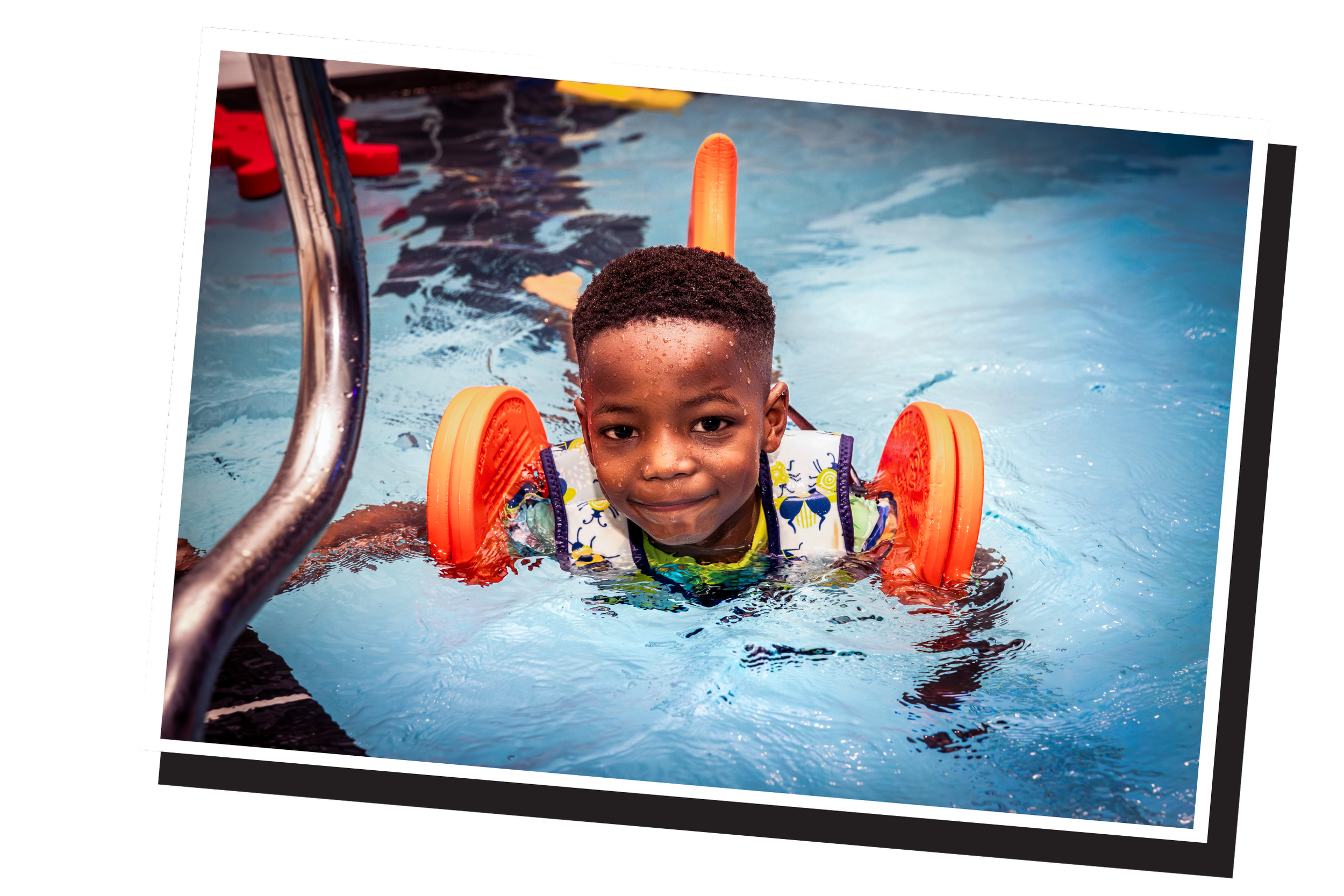Young child in a swimming pool with water and orange floaties on shoulders, smiling at the camera.