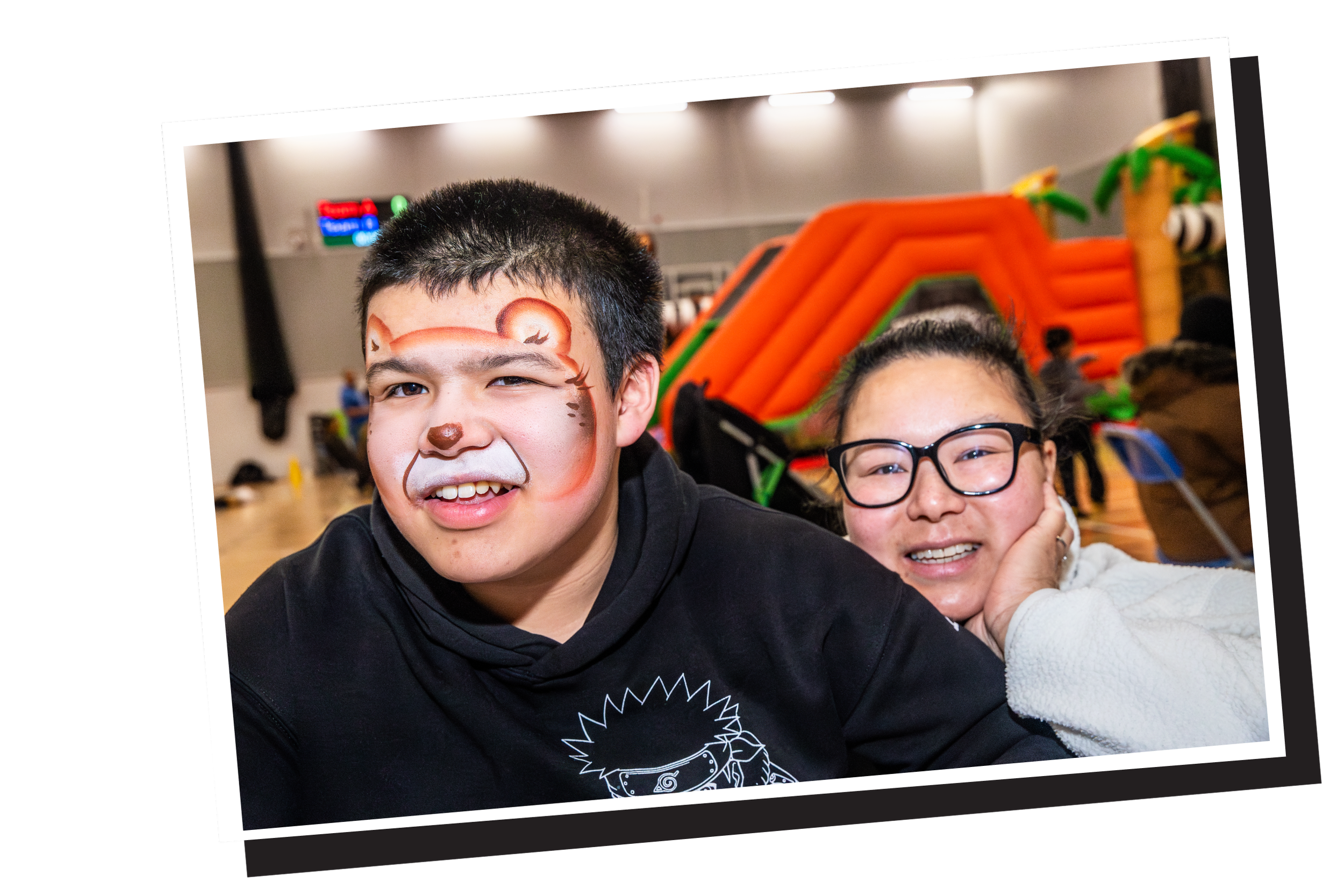 Two kids smiling, one with face paint of a bear and the other wearing glasses, in an indoor setting with inflatable structures in the background.
