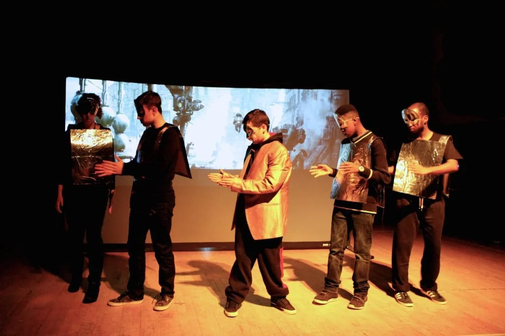 The image shows a group of young people on stage in costume appearing to be acting as robots or similar