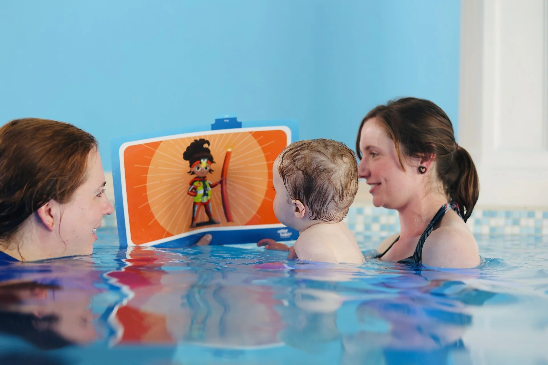 The image shows a baby in the pool with their parent and instructor looking at an orange image with a figure on it