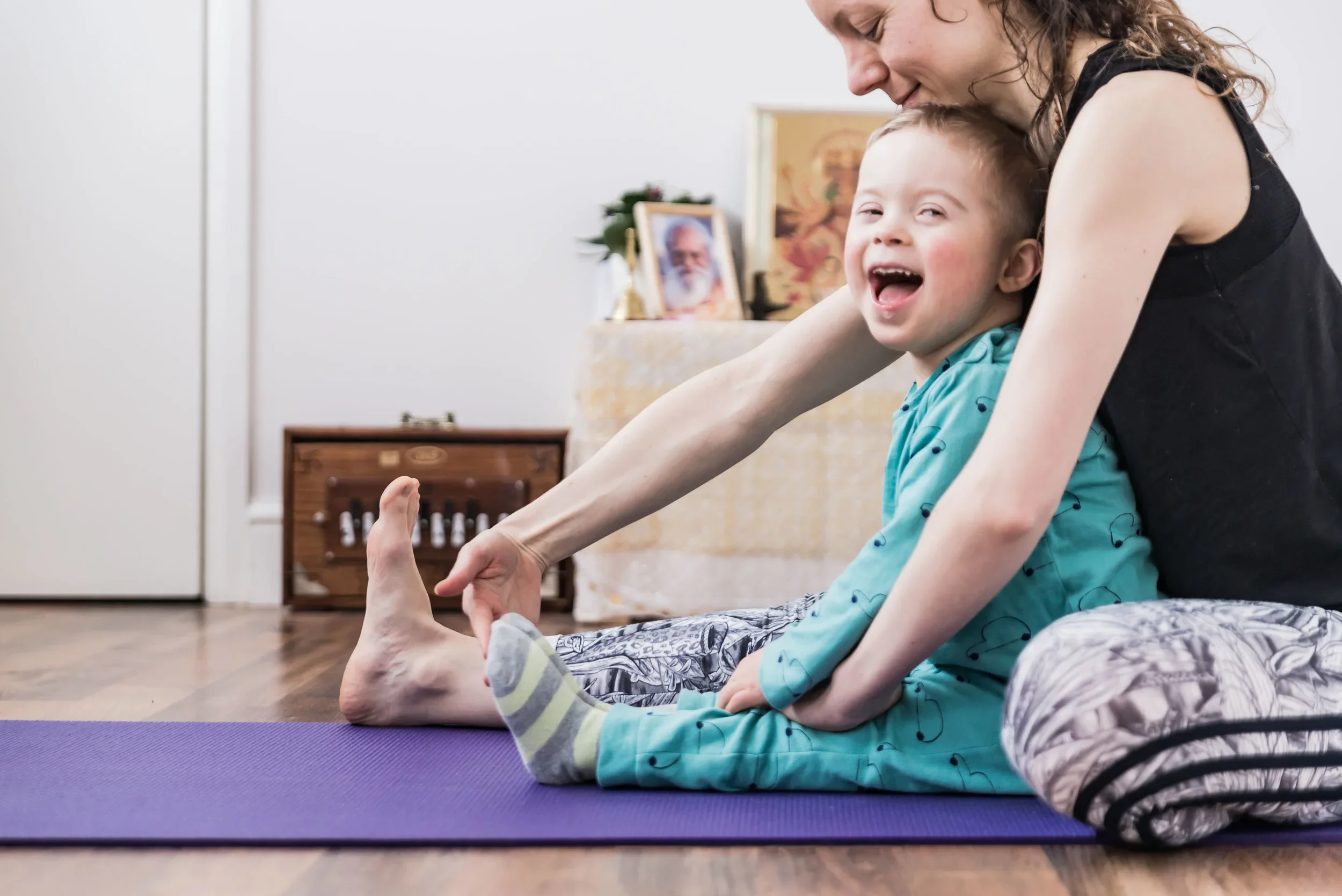 The image shows a young child engaging with stretches with help of the yoga teacher