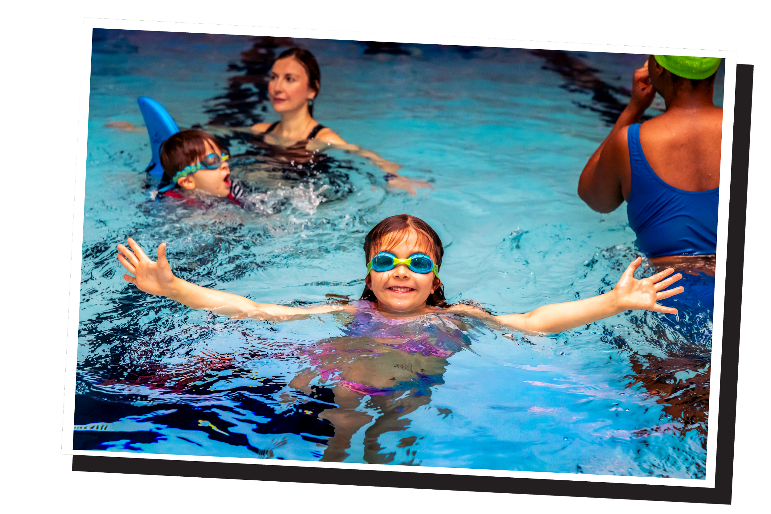 Children swimming in a pool with goggles, accompanied by adults, some wearing swim caps.